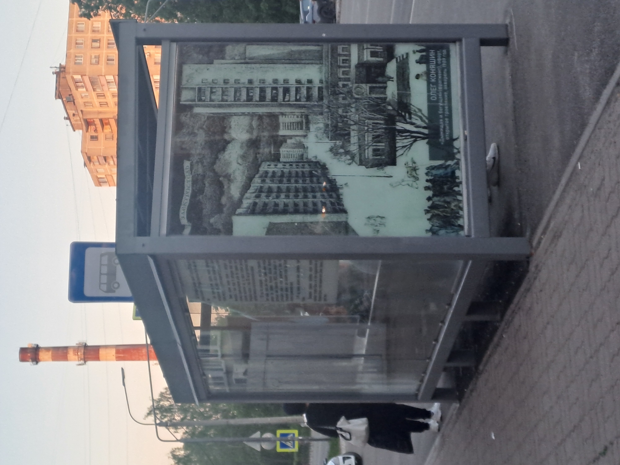 The image shows a bus stop in Elektrostal, Russia. The bus stop has a display case with a print of an etching by Oleg Konyashin, titled "Smoking and Running" (fragment), a 1989 etching, open etching, watercolor. A tall, orange and white striped industrial smokestack is visible in the background. In the reflection of the bus stop glass, apartment buildings are visible. A person wearing a black coat, white pants, and white shoes is standing by the bus stop. There is a street sign visible.