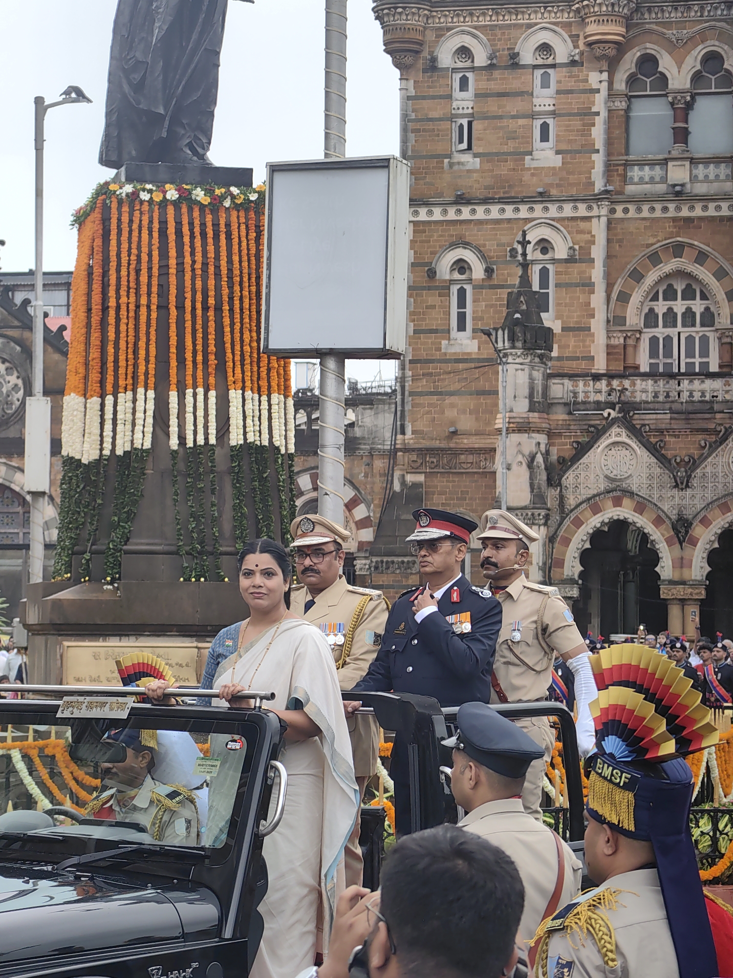 This image captures a formal event likely related to a patriotic celebration in front of the Chhatrapati Shivaji Maharaj Terminus (CSMT) in Mumbai, India. The iconic Victorian Gothic architecture of the building forms the backdrop. In the foreground, a woman in a white sari stands next to a man in a military uniform. They are positioned in front of a large statue decorated with orange and white garlands, possibly representing the Indian flag. Several other individuals in military and police uniforms are also visible, some standing and others in a black open-top vehicle. A man in the vehicle is seen driving. The overall atmosphere appears to be one of respect and ceremony.
