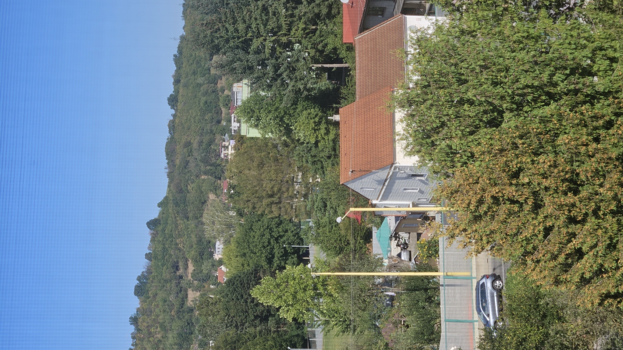 This is a high-angle, medium shot of a suburban area in Prešov, Slovakia. The scene is dominated by lush greenery and residential buildings under a clear blue sky. In the foreground, several trees with green and yellowing foliage are prominent. A silver SUV is captured in motion on a road bordered by a light-colored fence. Behind the road, a cluster of houses with red-tiled roofs are visible, along with a green umbrella and a motorcycle parked in a yard. Further in the background, a wooded hill rises, dotted with more houses and trees. The overall impression is one of a quiet, sunny day in a residential neighborhood surrounded by nature.