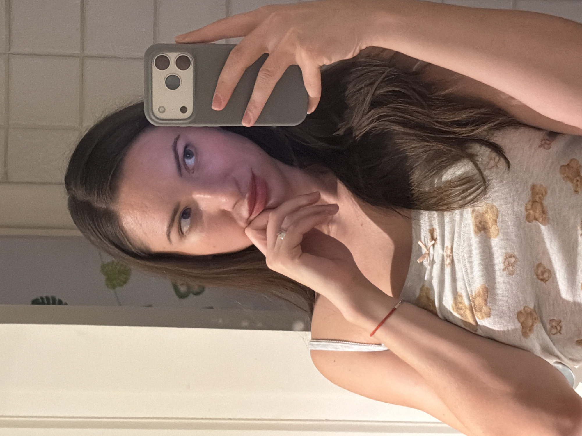A woman with long brown hair is taking a selfie in a bathroom. She is wearing a white camisole with a teddy bear print and a red string bracelet on her left wrist. Her right hand is holding a smartphone with a grey case. Her left hand is resting on her chin. The background shows a tiled wall and a doorway.
