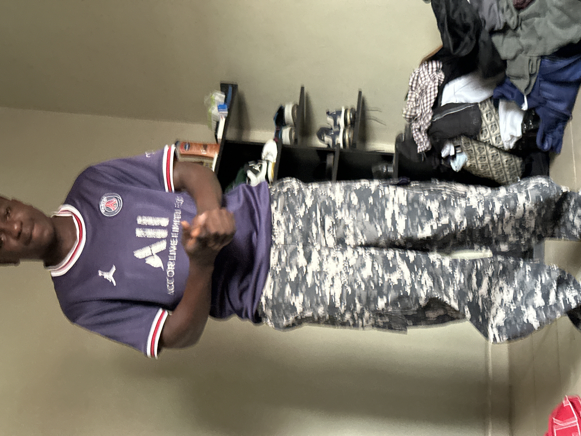 In an indoor environment, a young Black male stands facing forward. He is wearing a dark blue short-sleeved jersey with white and red trim around the collar and sleeves, featuring a white Jumpman logo and a circular crest. The jersey also has "AII" printed in white, followed by "ACCOR LIVE LIMITED" in smaller white text. His pants are camouflage in a digital pattern of black, grey, and white. His hands are clasped in front of his torso. To his right, a black shelf unit is visible, holding several pairs of shoes and some bottles on the top shelf. Below the shelves, a pile of clothes is scattered. The background wall is a muted, light-colored shade. The floor appears to be tiled or a similar hard surface. The lighting is diffuse and suggests an indoor setting.