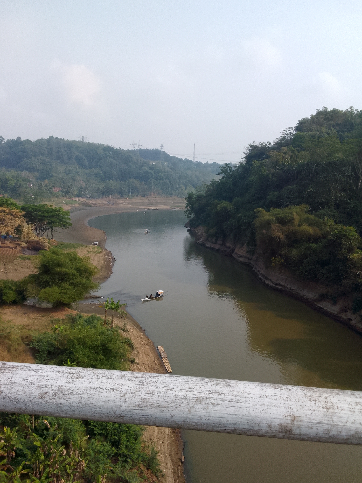 A wide, murky river carves through a lush, green valley. On the left bank, a sandy shore curves gently, dotted with sparse trees and some cultivated land. A small white boat with two people on board is navigating the river, heading away from the viewer. Further upriver, another boat can be seen in the distance. The right bank is steeper, densely covered with trees and vegetation, with some layered rock formations visible along the waterline. In the background, rolling hills covered in trees stretch towards a hazy sky. A white pipe, likely part of a railing or bridge, runs across the foreground of the image, partially obscuring the view of the river and banks. Power lines and towers are faintly visible on the distant hills, suggesting human infrastructure within the otherwise natural landscape. The overall impression is one of a serene, rural environment in a tropical region.