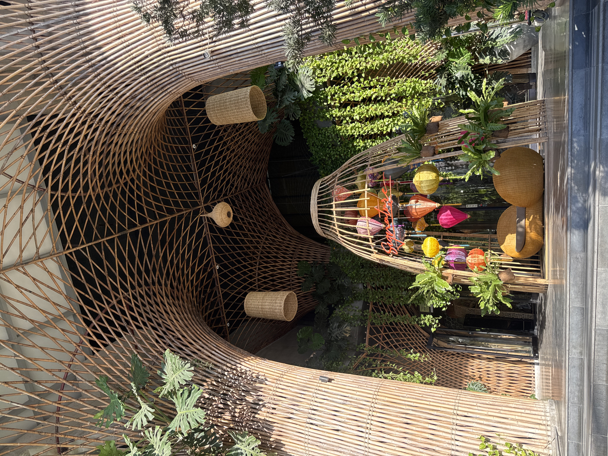 The image captures the exterior entrance of a restaurant in Nha Trang, Vietnam. The entrance features a distinctive arched canopy constructed from natural bamboo poles, creating a latticed effect overhead. Hanging from the bamboo structure are several cylindrical woven lampshades, adding to the rustic ambiance.
In the foreground, a prominent decorative element is a large, conical bamboo cage filled with colorful paper lanterns. These lanterns vary in shape and hue, including pink, purple, orange, and yellow, and some appear to be lit from within. Beside the cage, two rounded, woven rattan chairs with cushions are placed, suggesting a waiting or seating area.
The entrance is flanked by abundant greenery. Lush plants, including large-leafed philodendrons and trailing vines, cascade down the sides of the bamboo structure and form a living wall behind the central decorative elements. A glimpse into the restaurant through a glass door reveals more interior darkness and hints of architectural elements.
The ground in front of the entrance is paved with reflective tiles, and a set of grey steps leads up to the main entry area. The overall scene is bathed in natural sunlight, creating distinct shadows and highlighting the textures of the bamboo and foliage. The architectural style and the vibrant lanterns suggest an Asian-inspired, possibly Vietnamese, dining establishment.
