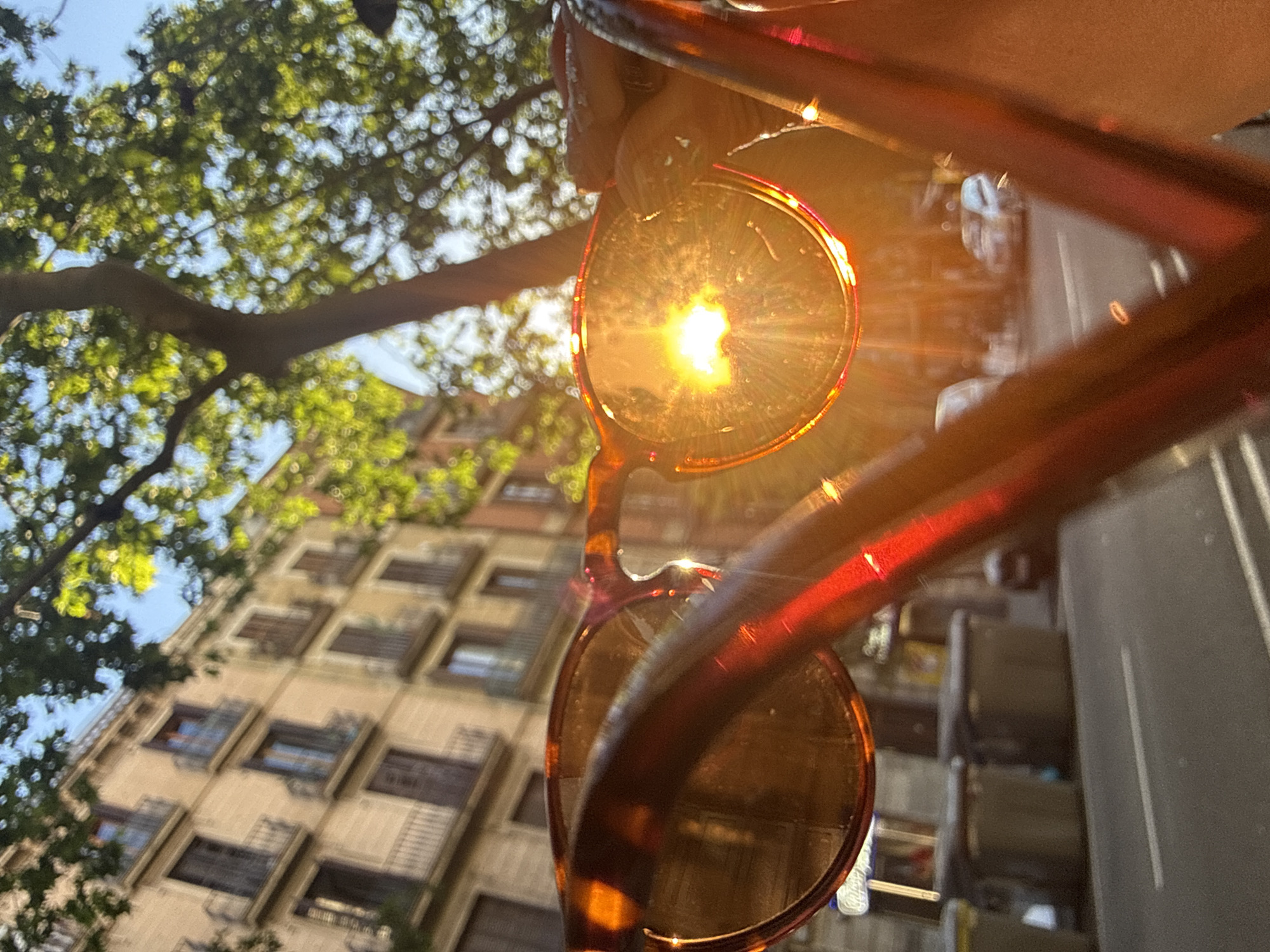 The image depicts a pair of sunglasses reflecting a bright sun and a blurry background of a building, trees, and what appears to be a street scene in the city of Barcelona, Spain. The building is multi-story with many windows. The tree has green leaves. The sunglasses have an amber-colored frame.