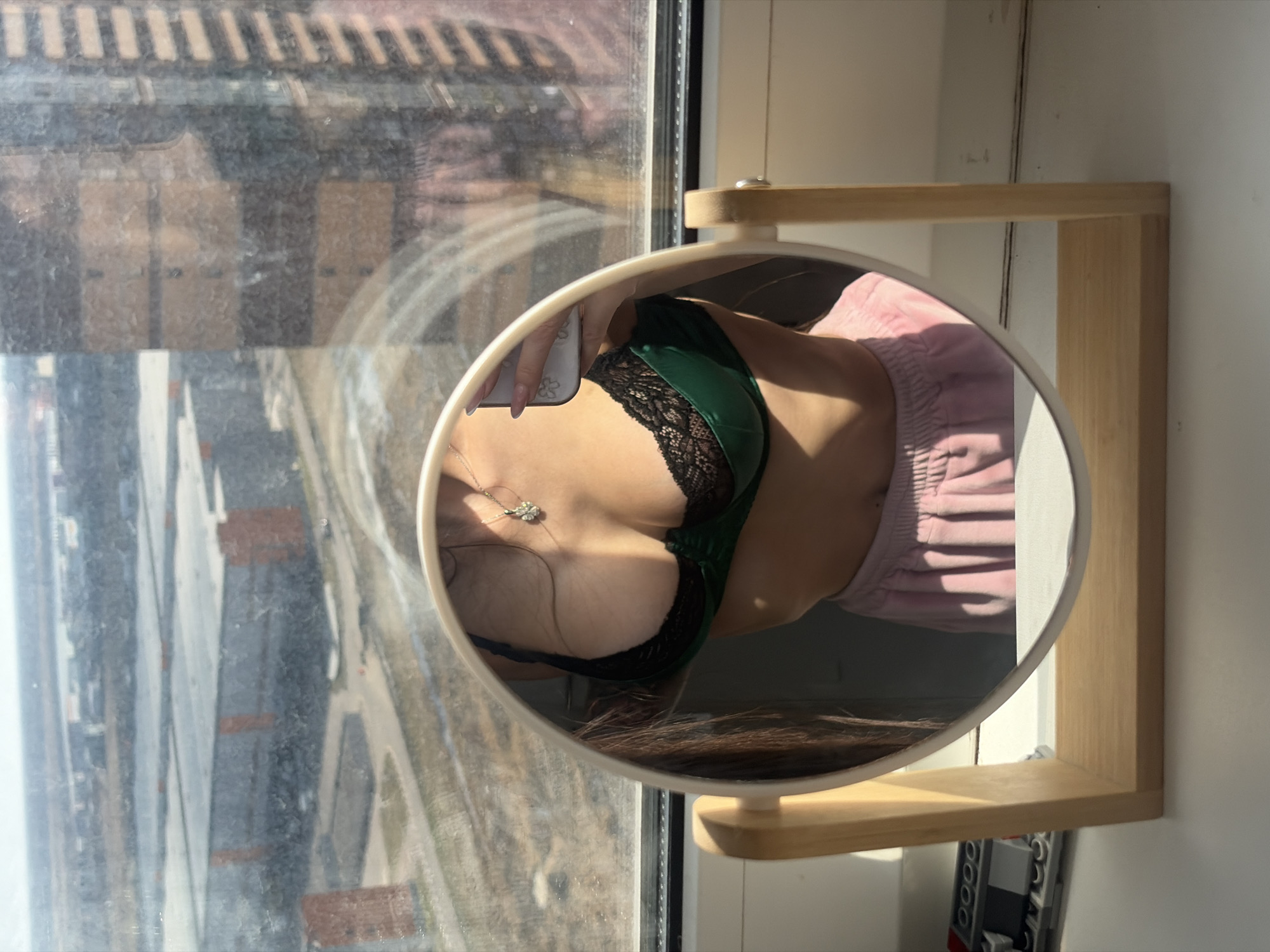 The image depicts a mirror selfie taken from a high-rise apartment in Saint Petersburg, Russia, overlooking an industrial area and the Gulf of Finland.
**Scene:**
A white oval desktop mirror with a light wooden stand is centered on a sunlit white window sill. Through the slightly dirty window glass, a hazy urban-industrial landscape is visible under a bright, clear sky.
**People:**
The upper torso of an individual is reflected in the mirror. The person is wearing a dark green bra with black lace trim and light pink velvet shorts or sweatpants. A silver necklace with a multi-part pendant hangs on their chest. The individual's left hand, with manicured light pink nails, holds a white smartphone with a floral-patterned case, angled for a mirror selfie. Long brown hair is visible along the left side of the reflection.
**Objects and Environment:**
* **Interior:** The white window sill supports the mirror. To the left of the mirror stand, a small collection of Lego bricks (grey, black, white) is visible. Strong sunlight casts shadows on the sill.
* **Exterior:** The view through the window reveals large, modern industrial or warehouse buildings with flat roofs and distinct grey facades featuring reddish-brown vertical panels. Dirt paths or unpaved areas are visible between these structures. In the far distance, a wide, hazy body of water, identified as the Gulf of Finland, meets the horizon. A tall, multi-story building with numerous horizontal bands, likely another high-rise, is reflected in the upper right portion of the window glass.
**Location in Saint Petersburg:**
Based on the combination of large, modern industrial/warehouse complexes, proximity to the Gulf of Finland, and the view from a high-rise residential building, the location is identified as the **Krasnoselsky District** in the southwestern part of Saint Petersburg. This district features numerous modern residential developments bordering industrial zones with views towards the Gulf.