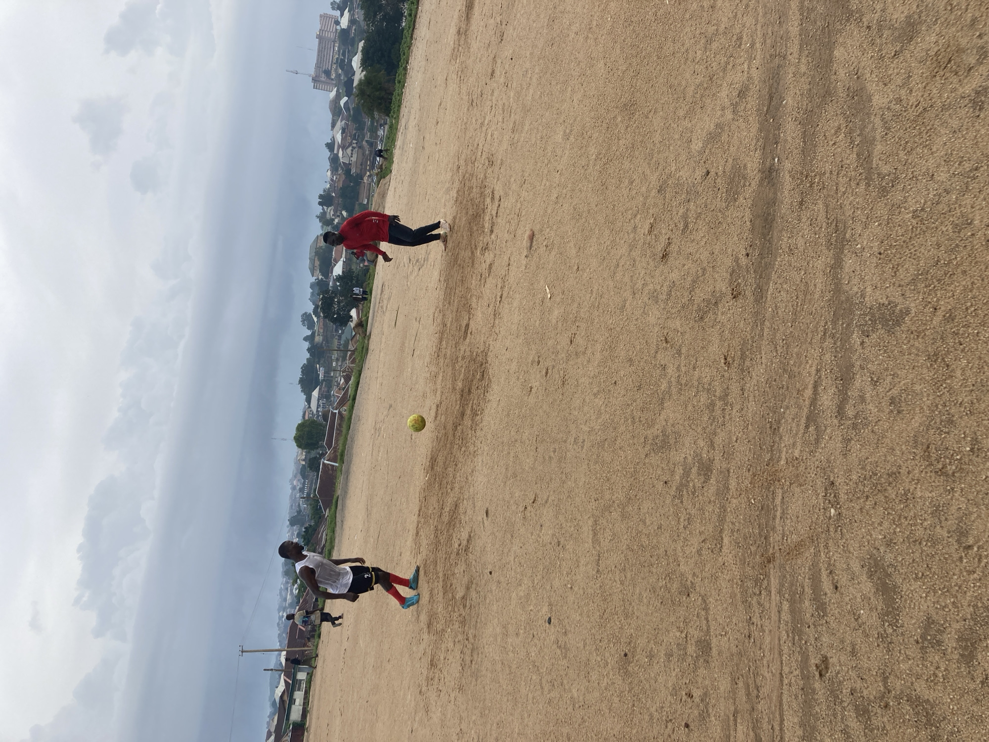 In the city of Jos, Nigeria, on a dusty, uneven dirt field, two young men are engaged in a game of soccer. The man on the left, wearing a white tank top, black shorts with red accents, and red socks, is positioned to kick a yellow soccer ball that hovers in mid-air. His opponent, on the right, is dressed in a red long-sleeved shirt and dark pants. The background reveals a cityscape with numerous houses and buildings, a large hotel with a crane nearby, and a cloudy sky. The overall scene depicts an informal soccer match taking place in a suburban or semi-urban area of Jos.