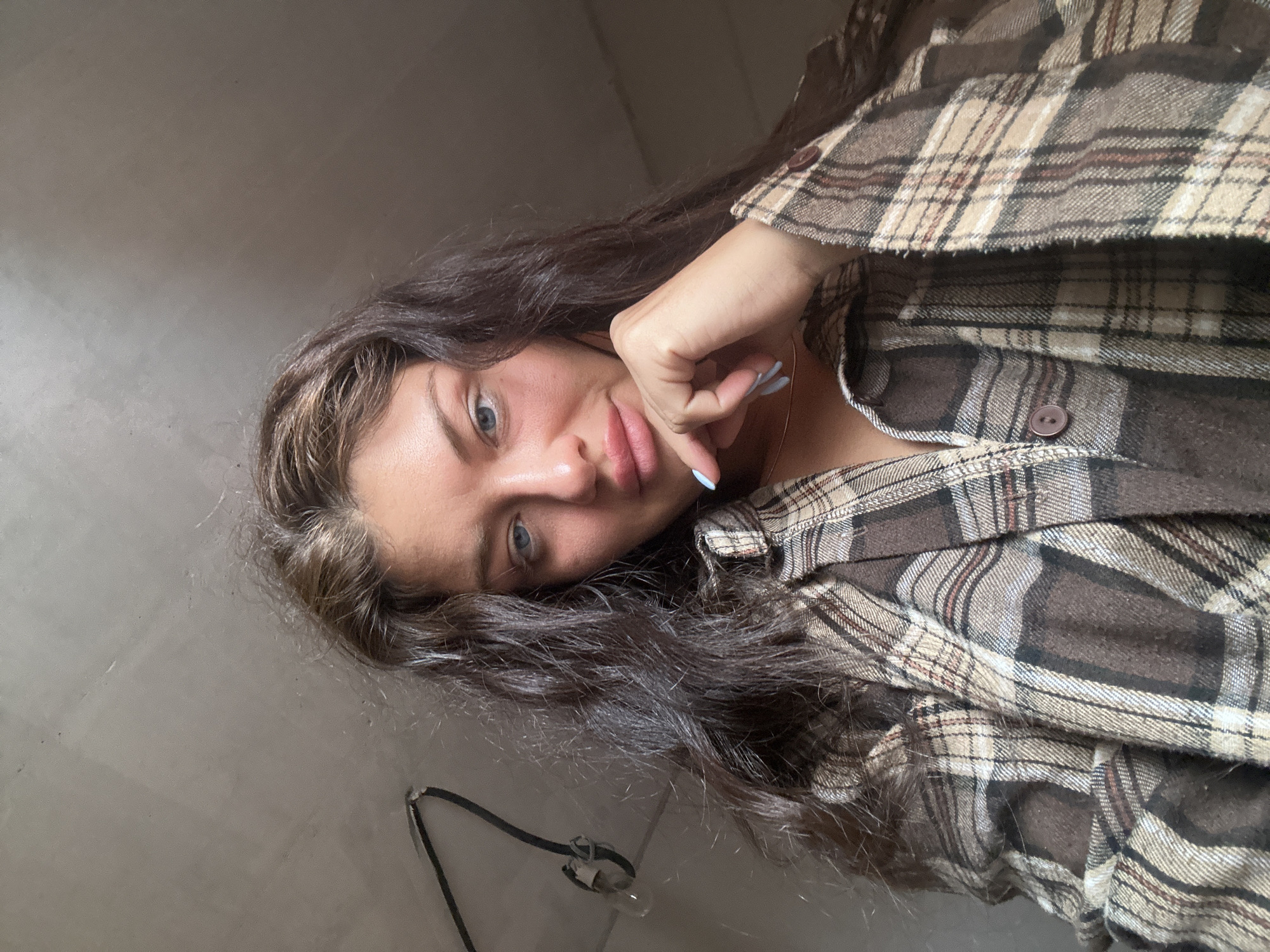 A young woman with long, dark hair and light eyes is shown in a close-up, slightly angled selfie. She is wearing a brown and tan plaid flannel shirt, partially unbuttoned. Her left hand is resting near her face, and her fingernails appear to be painted light blue. A thin gold necklace is visible around her neck. To the left of her head and below, part of a wall is visible, and a pair of glasses or a lanyard hangs down near the wall. The location is listed as the city of Unknown, Unknown.