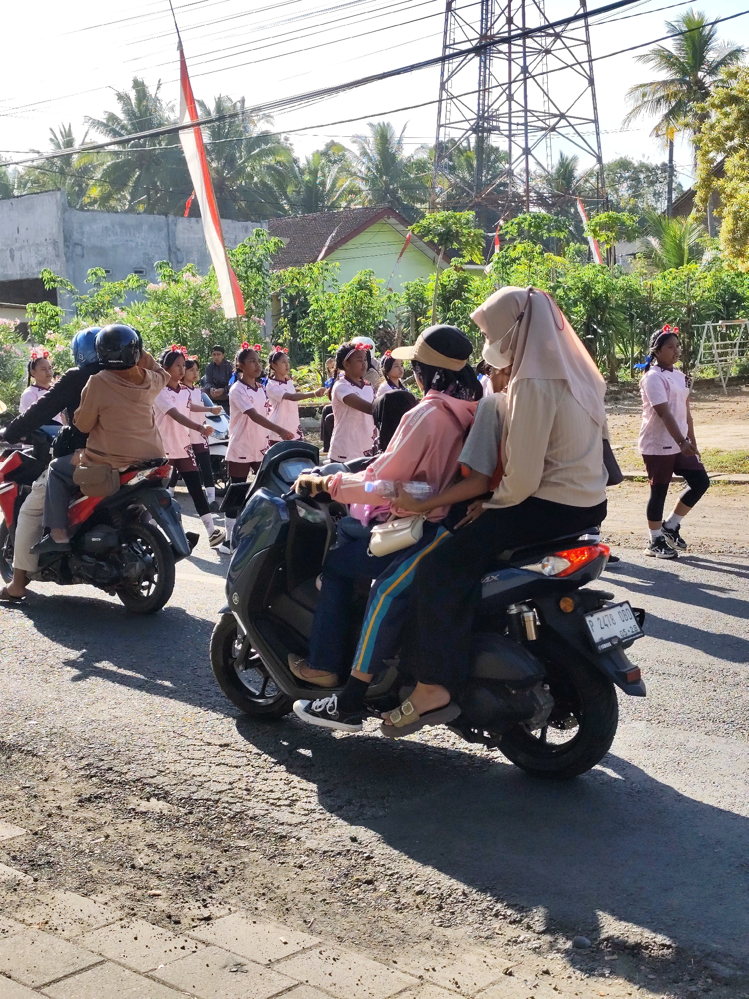 The image depicts a street scene in Tegalarum, Indonesia, likely during a local celebration or parade. In the foreground, two motorcycles are prominently featured. The motorcycle on the right is dark colored, with a license plate reading "P 2478 OBD". Two individuals are seated on this motorcycle; the front rider is wearing a light brown hijab and a mask, while the passenger behind wears a pink long-sleeved shirt with stripes and dark pants. The motorcycle on the left is red and black, carrying two passengers. The rider is wearing a dark helmet and a light brown top, and the passenger behind is wearing a light brown top and dark pants, also with a helmet.
Behind the motorcycles, a group of young girls, appearing to be students, are walking in formation. They are dressed in light pink uniforms with red headbands. Some are looking ahead, while others are engaged with their surroundings. Further in the background, more individuals are visible, including adults and possibly more children. A tall metal tower, resembling a telecommunications tower, stands in the upper right portion of the image. Indonesian flags, red and white, are visible in the background, tied to poles and draped between structures, suggesting a patriotic event. The environment includes trees, houses with red-tiled roofs, and power lines overhead, all bathed in bright sunlight, casting shadows on the asphalt road. The road surface is a mix of asphalt and gravel, with a paved sidewalk visible on the left.