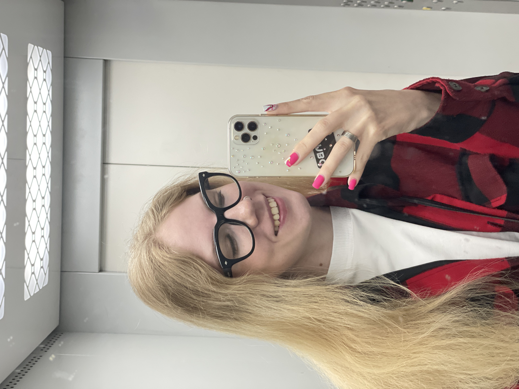 A young woman with long blonde hair smiles while taking a mirror selfie indoors. She is seen from the chest up, looking directly at the camera with a happy expression. She wears black-rimmed glasses, and a subtle nose ring is visible. Her attire includes a red and black plaid shirt worn over a white crew-neck undershirt.
Her left hand holds a light-colored smartphone, likely an iPhone given the prominent triple camera setup, which is encased in a clear cover embellished with numerous small, reflective rhinestones or gems. Her fingernails are painted a bright, vibrant pink, with some fingers featuring white and red abstract designs. A metallic ring is worn on her left ring finger. A black sticker with white text, possibly reading "JBC," is affixed to the back of the phone case.
The background consists of plain, light-colored walls, possibly white or light gray, suggesting an indoor environment such as a corridor or elevator. On the far left, a vertical panel with a distinct diamond-shaped lattice pattern is visible, which could be a decorative element or part of a ventilation system. Horizontal lines or seams are present on the wall surfaces, indicative of paneling or construction. The overall scene conveys a casual and cheerful moment.