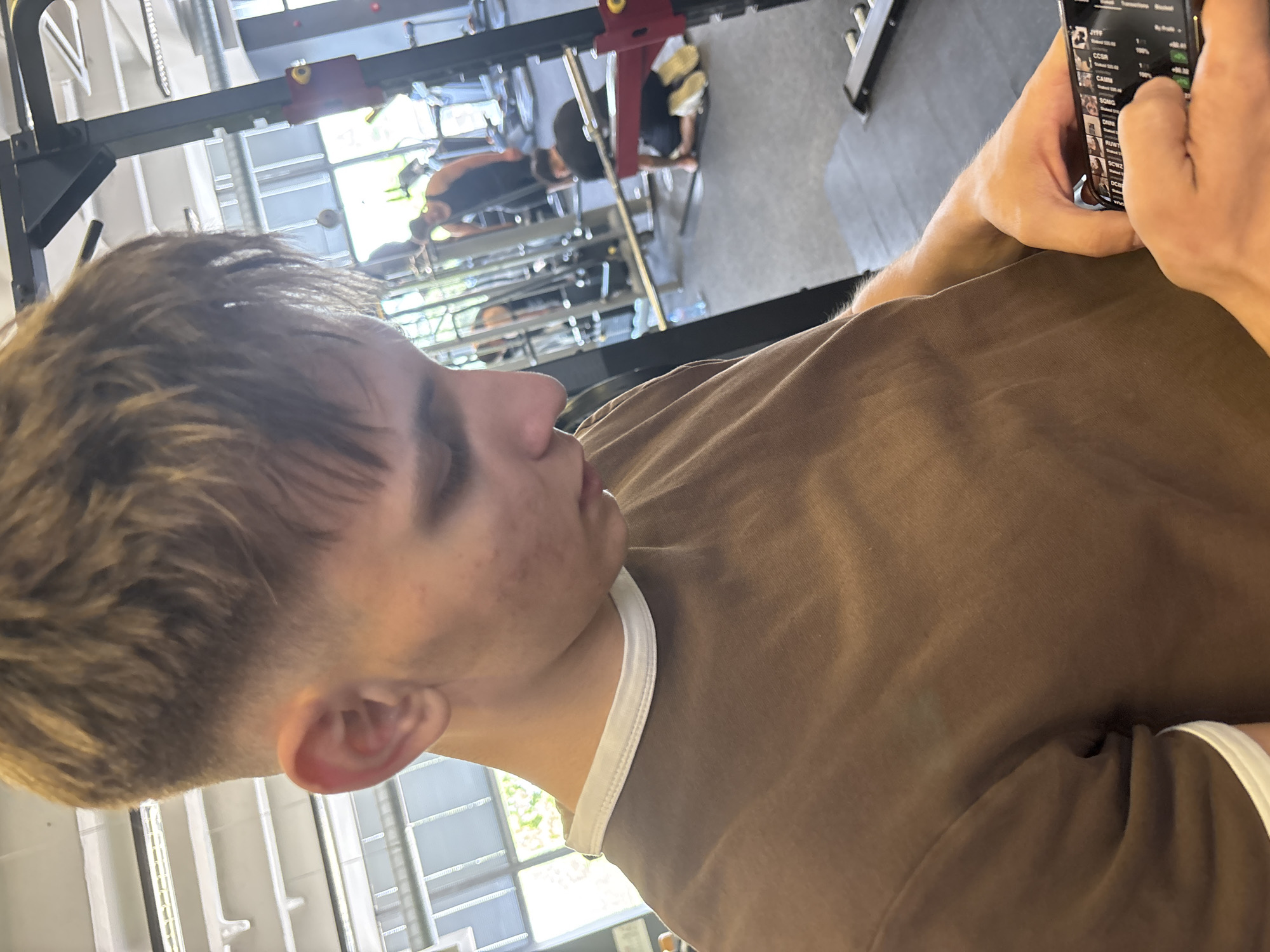 The image captures a young person in the foreground, seen from the side, looking intently at a smartphone held in their right hand. The person has short, light brown or blonde hair with a faded cut, and is wearing a plain brown short-sleeved t-shirt with a white collar lining. Their left ear is visible, and there are some minor blemishes on their skin.
The smartphone screen displays what appears to be a financial or stock market application. Visible text includes headings like "Transactions" and "Blocked" at the top. Below, a list of abbreviations or stock tickers is shown, such as "JTF", "CCSR", "CAIM", "SGMG", "GWZZ", "DHRR", "RUWT", "GCW2", "GCBS", and "OCB". Next to these, percentages and numerical values are visible, for example, "100%" and "+00.00".
The background is an indoor gym or fitness center, brightly lit by natural light streaming through large windows, indicating daytime. Various pieces of gym equipment are visible, including what looks like a power rack or squat rack, and other exercise machines. Several other individuals are present in the blurred background, some appearing to be exercising or engaging with the equipment. The overall setting is modern and well-lit.