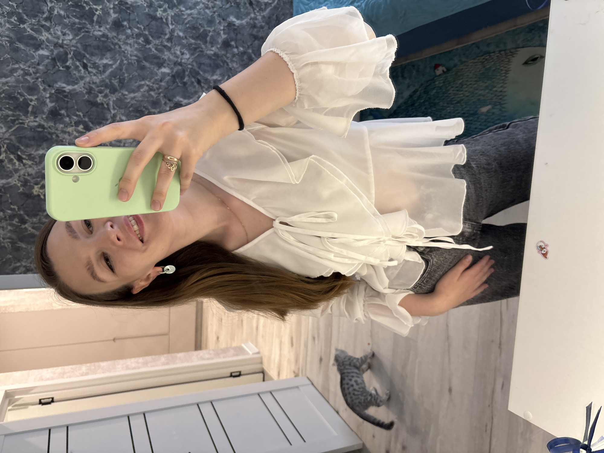 A young woman is taking a selfie in a well-lit interior. She is wearing a white ruffled blouse and dark jeans. A small spotted cat is walking away from the camera in the background. The environment appears to be a residential hallway, possibly in a modern apartment building. There is a door with horizontal white panels on the left and a marbled pattern on the wall behind the woman. The floor is light-colored laminate. The overall impression is of a casual, domestic moment captured through a smartphone camera. The image was taken in Izhevsk, Russia.