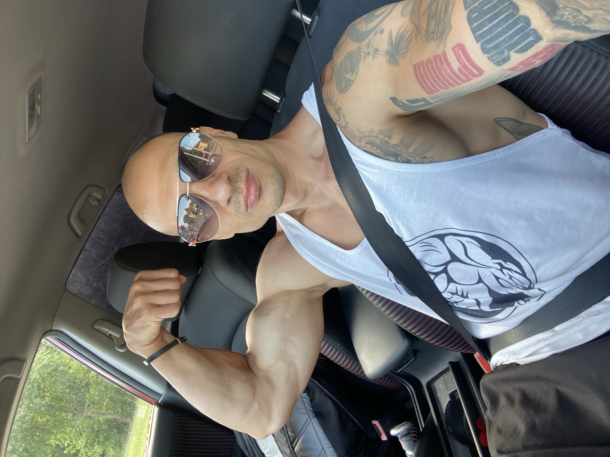 A bald, muscular man with sunglasses is sitting in the driver's seat of a car. He is wearing a white tank top with a graphic design and a black bracelet on his left wrist. His left arm is flexed, showcasing his bicep. He has tattoos on his right arm. The interior of the car is visible, including the dashboard, seats, and a seatbelt. The background outside the car appears to be a natural setting with trees. The image was taken in the city of Pleven, Bulgaria.