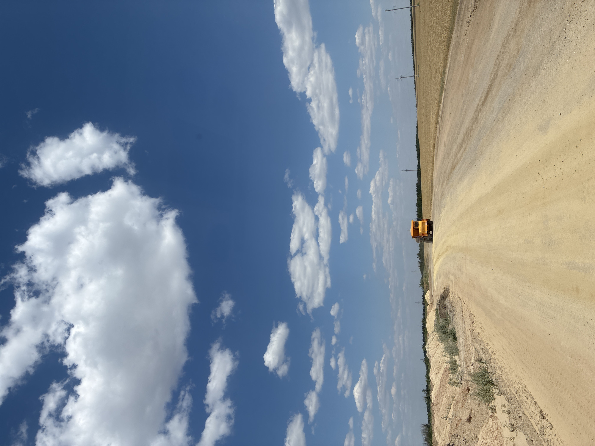 The image depicts a rural road in the city of Zhirnov, Russia, under a clear blue sky dotted with fluffy white clouds. A yellow construction vehicle is visible in the distance on the dusty, dirt road, which curves through a landscape of arid fields and piles of sand. The scene is characteristic of a construction or agricultural area in a dry climate.