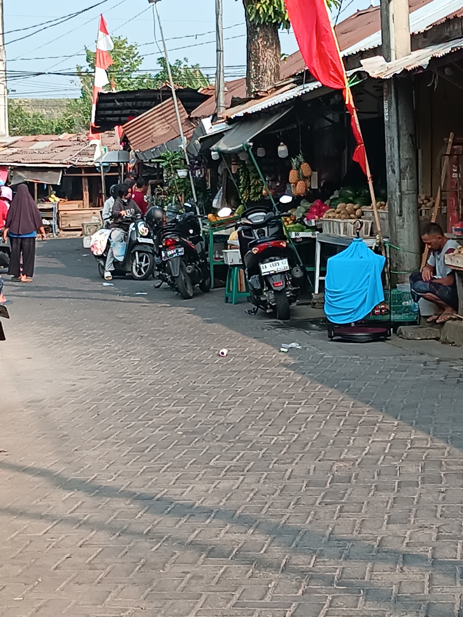 The image depicts a street market scene in Yogyakarta, Indonesia, possibly on a sunny day given the shadows. In the foreground, a paved street with interlocking brick patterns is visible. To the left, a woman wearing a dark hijab and black pants walks away from the camera. Further down the street, two motorcycles are parked, one in front of the other. The front motorcycle is dark-colored with a rider wearing a gray hoodie and blue jeans. The rear motorcycle is also dark-colored.
The background reveals a row of market stalls with corrugated metal roofs. Some stalls are laden with fruits, including pineapples, bananas, and what appear to be watermelons or cantaloupes. Other stalls display other produce. Red and white flags, possibly Indonesian flags, are prominently displayed, suggesting a national holiday or event. A man in a light-colored shirt and dark pants is seated on the curb to the right, near a birdcage covered with a bright blue cloth. The environment appears to be a bustling urban street market.