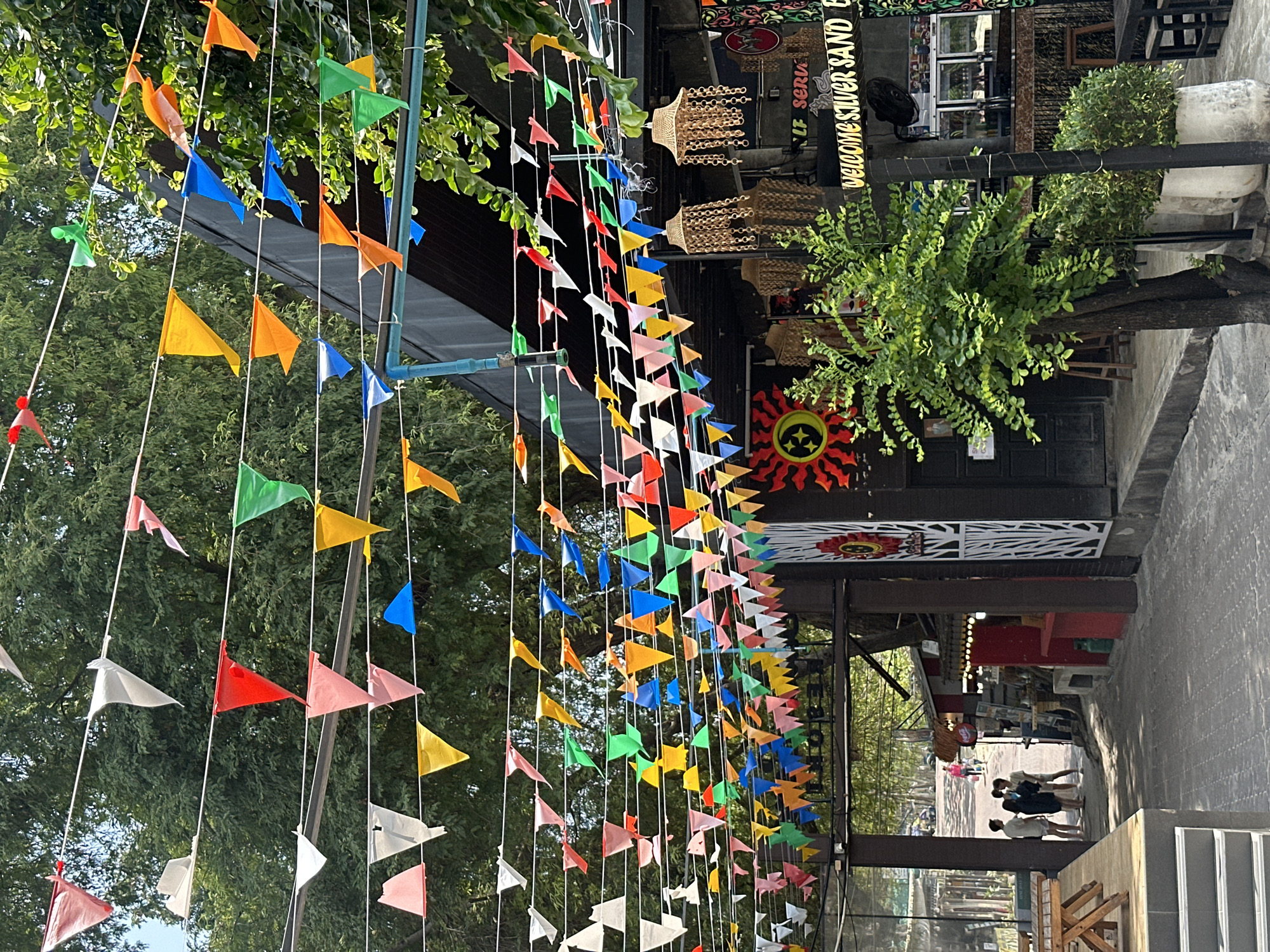 The image shows an outdoor scene in Ao Noi Na, Thailand, likely a street view. A vibrant display of colorful triangular flags hangs overhead, strung on white cords, creating a festive or celebratory atmosphere. The flags are various colors, including orange, yellow, green, blue, red, pink, and white.
On the right side of the frame, storefronts with wooden structures and dark-colored roofs are visible. Signs above the shops display words like "WELCOME SILVER SAND" in English. There are bamboo decorations hanging above the storefronts.
Several people are standing near the back of the storefront, near the end of the street. The street itself is light gray or concrete and appears to have some pedestrian activity. The scene has a sunny and bright ambiance. The environment includes elements of trees and greenery.