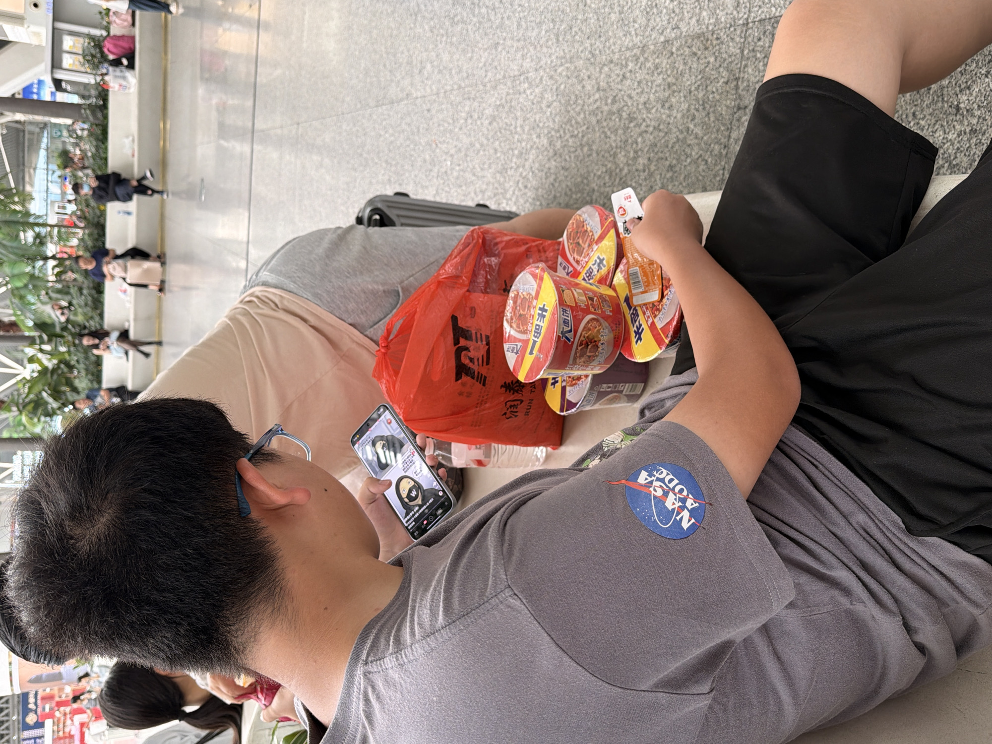 The image shows a close-up view of a young person sitting in an indoor waiting area, possibly at a train station or airport in Panyu District, China. The individual is wearing a grey t-shirt with a NASA "meatball" logo patch on the left sleeve and black shorts. They are holding a smartphone in their right hand, displaying a video or image with Chinese text. In front of them are several cups of instant noodles and a red plastic bag with Chinese characters. Behind the person, other individuals can be seen sitting on a bench in the background, some with luggage. The environment appears to be a clean, modern public space with tiled flooring and decorative plants.