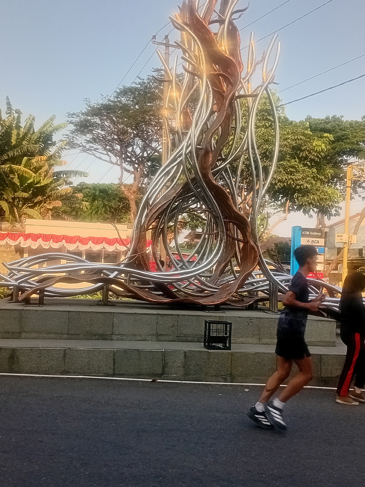 The image depicts a street scene in Bacin, Indonesia, featuring a large, abstract sculpture made of metallic and possibly wooden elements. The sculpture has a central, flame-like structure with intertwined, curved metallic tubes extending outwards, some appearing to flow along the ground. In the foreground, two individuals are jogging on an asphalt road. The person closer to the viewer, on the left, is wearing a dark t-shirt, dark shorts, and athletic shoes with white stripes. The person on the right is wearing dark pants with a red stripe and lighter-colored shoes. Behind the sculpture, a building with a red and white decorative trim is visible, along with trees and utility poles. A blue sign that says "ATM Gallery BCA" is also visible. The sky is clear and blue, suggesting it is daytime, possibly late afternoon due to the lighting. A black plastic crate is placed on a stone step near the sculpture.