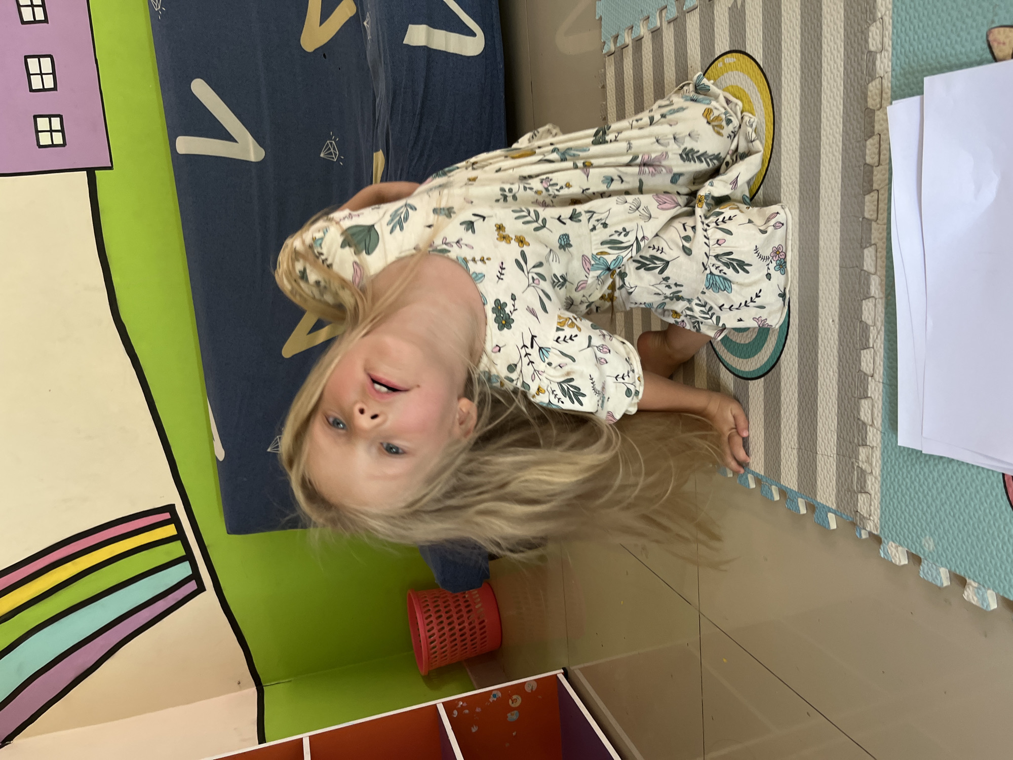 A young girl with long blonde hair and blue eyes is captured in an upside-down, close-up shot. She is wearing a white dress with a floral pattern of blue, green, and pink flowers and leaves. Her head is tilted back, and her mouth is slightly open as if she is laughing or speaking.
The setting appears to be indoors, possibly a playroom or classroom, with colorful decorations. To the left, a section of a wall mural features a purple building with white-outlined windows and a bright green stripe. A rainbow mural with purple, blue, green, yellow, and orange stripes is also visible on the left side.
The floor is covered with interlocking foam mats in alternating stripes of gray and light blue. A pink mesh basket is placed on the floor. To the right, a stack of white papers rests on the mats. In the background, a dark blue fabric with white "V" shapes and diamond patterns can be seen.
The lighting suggests it is daytime, but the lack of visible windows makes it difficult to determine the exact time or weather. The girl's expression conveys a sense of playfulness and joy. The image is taken in Kuta, Indonesia, suggesting a tropical or warm climate. No visible text is present in the image.