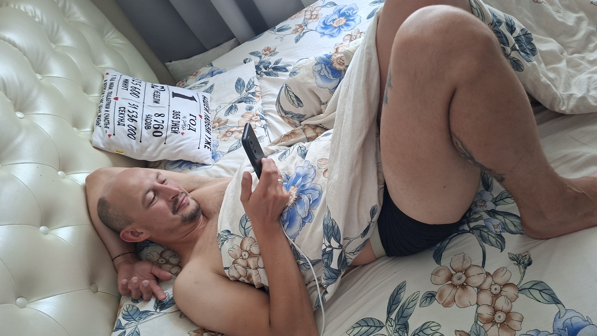 A man is lying in a bed, partially covered by a blanket with a floral pattern. He is looking at his phone, which he holds in his right hand. The man has a shaved head and a goatee. He is wearing black underwear. In the background, a pillow with text is visible. The bed has a large, tufted headboard. The scene appears to be indoors, in a bedroom. There is no information in the image that would allow for a specific location in the city of Myadzyel, Belarus.