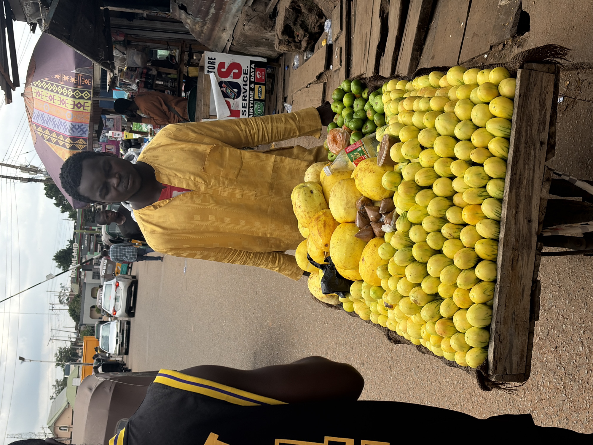 This image captures a street scene in Jos, Nigeria, likely near a market or busy intersection. A man in a yellow traditional garment stands behind a cart laden with an abundance of yellow fruits, possibly local varieties of mangoes or papayas, and smaller green fruits, which appear to be limes or small gourds. The cart is positioned on a paved road, with other vehicles and pedestrians visible in the background, suggesting an active urban environment. To the right of the fruit cart, a makeshift stall displays various items, including a bottle of Coca-Cola and what appear to be mobile phone services advertisements. The overall atmosphere is that of a typical Nigerian street market, with commerce and daily life unfolding.