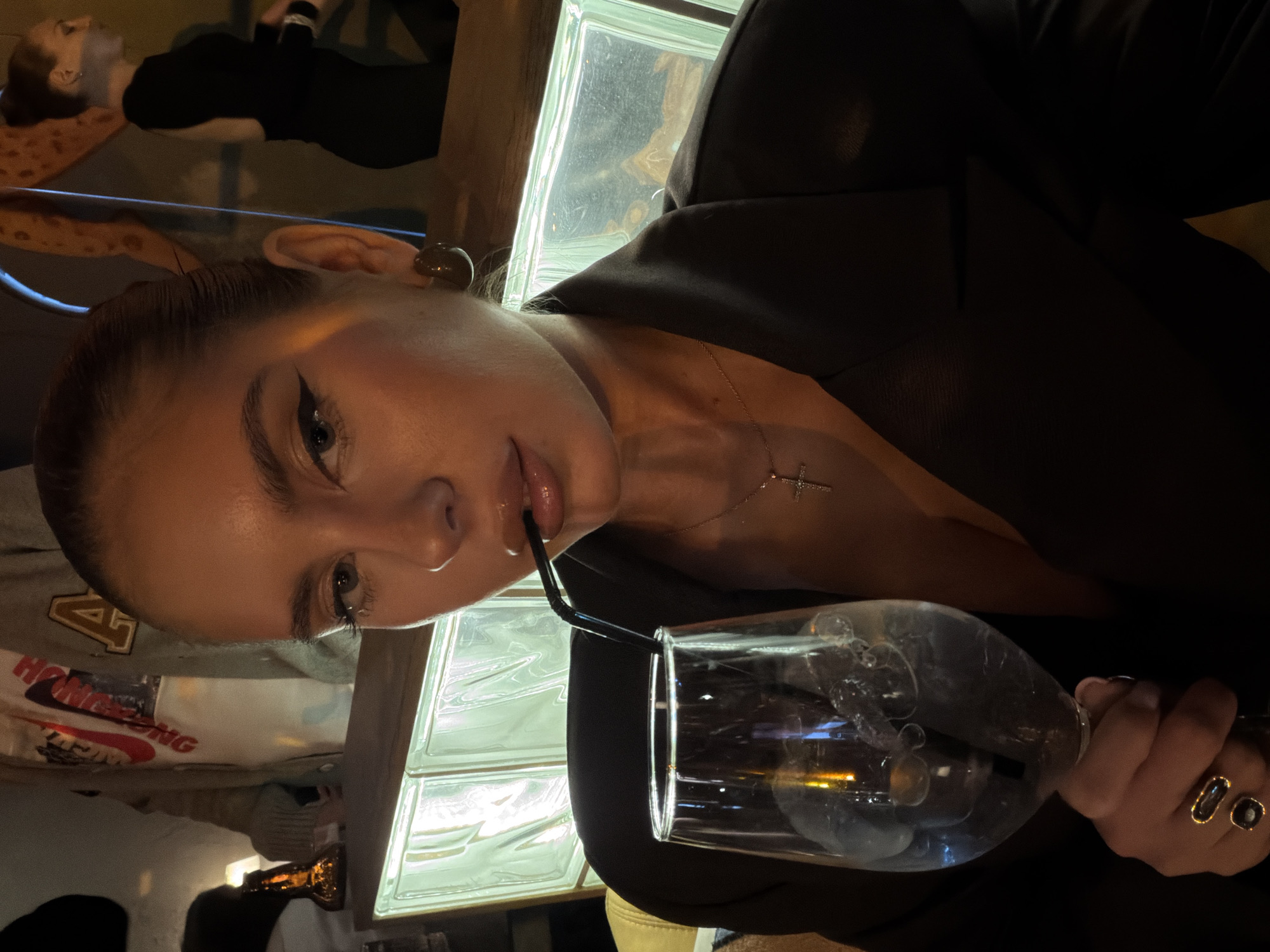 The image shows a woman in a dark jacket, holding a glass with a straw. She has brown hair pulled back, earrings, and a necklace with a cross. She is holding the glass with her right hand, which has rings on the fingers. The background contains a wall of glass blocks and other people in the periphery. I cannot determine a location in the city of Krasnoyarsk, Russia.