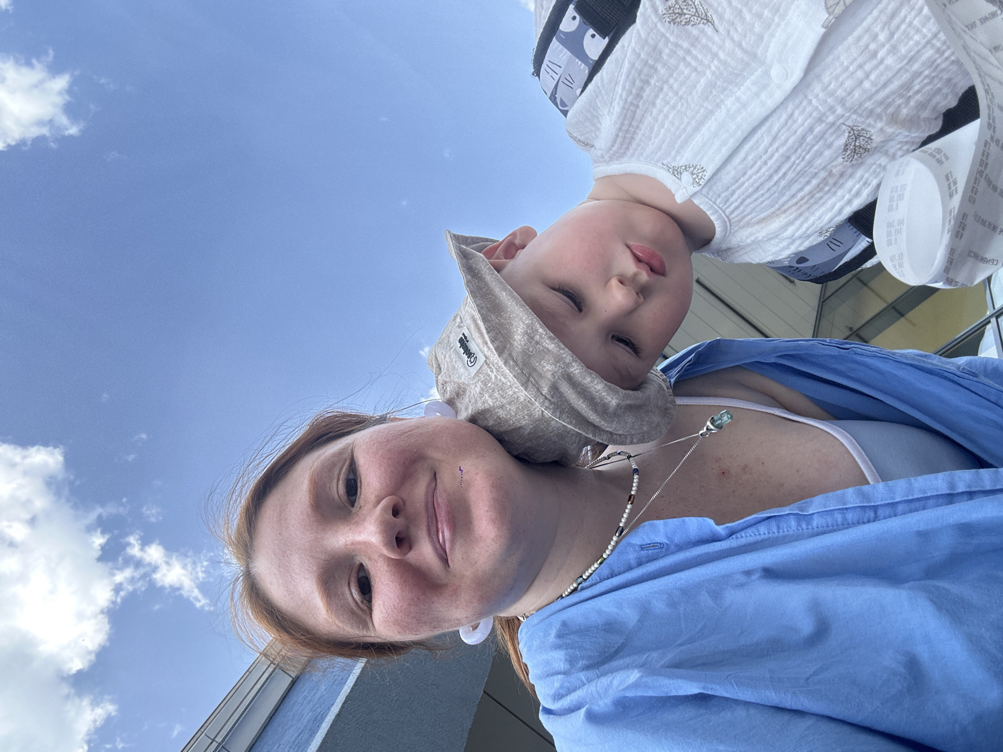 A medium shot captures a person, a woman with fair skin and reddish hair, positioned below and behind a child with a tan sun hat. The child is leaning into the frame and appears to be in a baby carrier. They are both looking downwards at the camera. The woman is wearing a light blue button-up shirt and a necklace. The background shows a partially cloudy blue sky and the corner of a modern building with blue and white elements. Based on the architectural style of the visible building and the type of baby carrier, the photograph might have been taken in an urban setting, but specific details to pinpoint the exact location in Nizhny Novgorod, Russia, are missing.