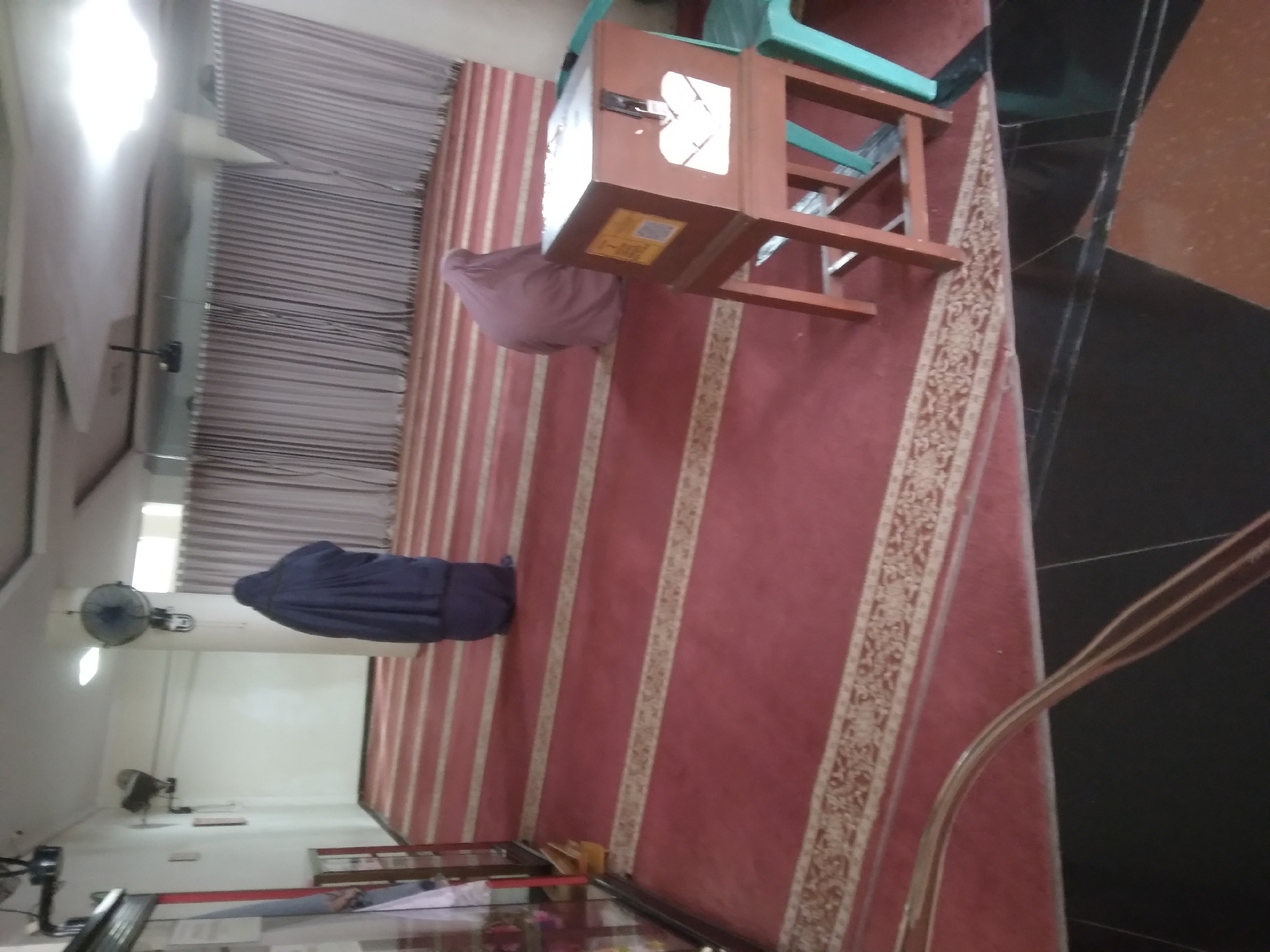 In a room with red carpet and white striped patterns, two individuals are seen praying. The room has white walls and a white ceiling with a light fixture. The environment appears to be indoors, possibly a place of worship given the prayer mats and the presence of two individuals in prayer attire. The furniture includes a wooden box on a stand, and a green chair. The lighting is adequate. The image suggests a serene and solemn atmosphere.