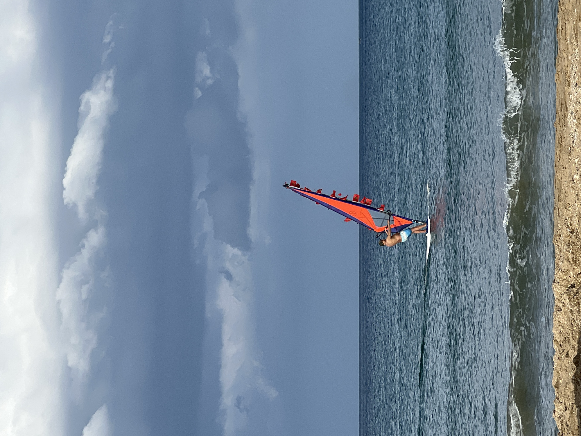 The image shows a man windsurfing on the Mediterranean Sea near M'Diq, Morocco. The man is wearing blue shorts and has his back to the viewer. He is standing on a white windsurf board, holding onto the boom attached to a large orange and blue sail. The sail is angled upward, suggesting the wind is propelling him across the water. To the right is the coastline with a narrow sandy beach. The sea is a deep blue color, with small waves lapping the shore. In the background there is a blue sky with dark clouds.