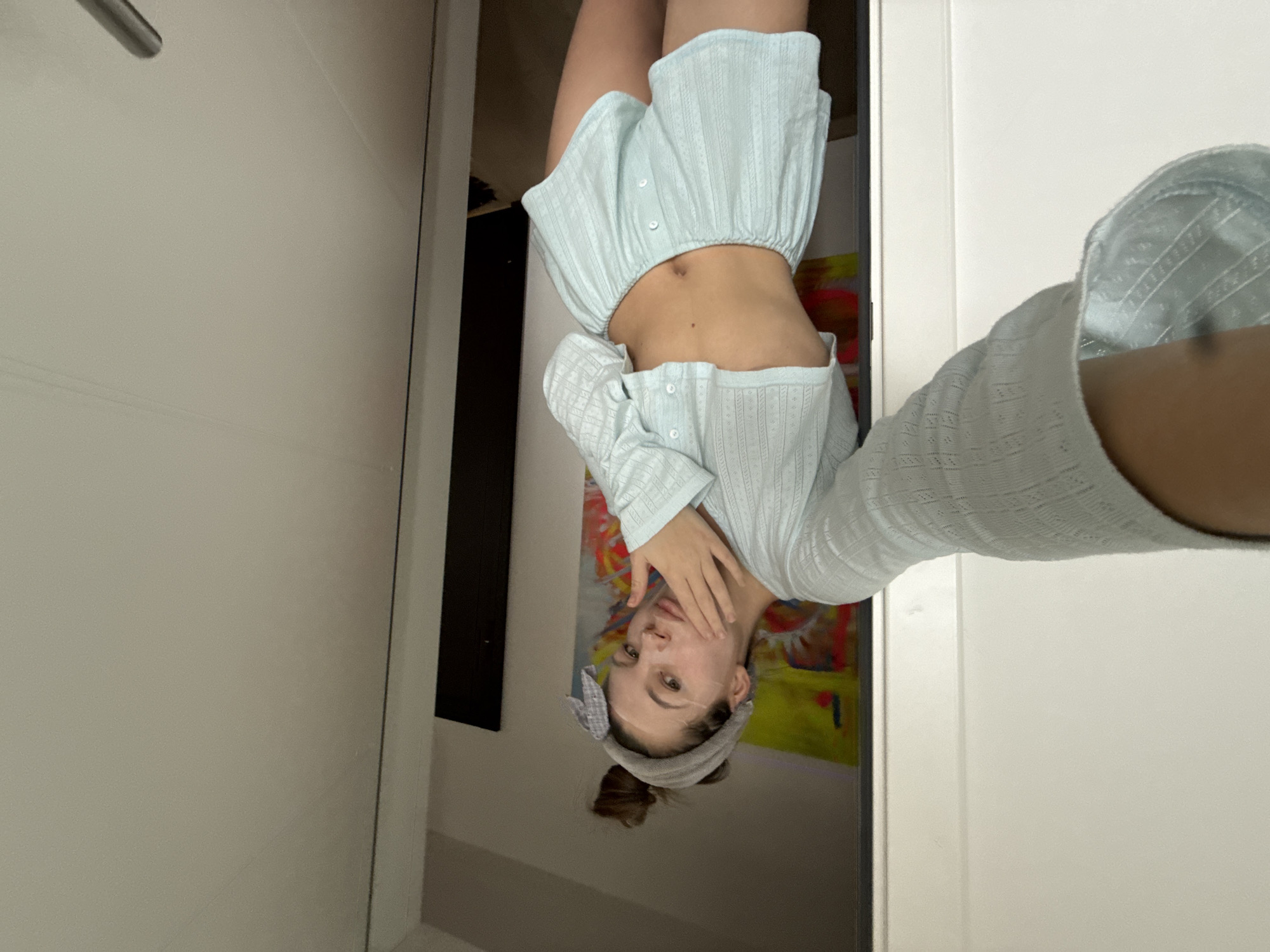 The image displays a female subject indoors, within a residential setting in Vozdovac Urban Municipality, Serbia. The subject, a young adult, is centered in a doorway or against a doorframe, capturing a self-portrait. Her brown hair is styled in an updo and secured by a grey fabric headband with a bow. A translucent facial mask is visible on her face. She is dressed in a light blue, textured two-piece loungewear set, comprising a cropped, long-sleeved, button-front top that exposes her midriff, and matching high-waisted shorts with visible front buttons and an elasticated waistband. Her right arm is extended to hold the camera, with her forearm partially visible in the lower-left of the frame. Her left hand's fingers are positioned against her right cheek and chin. Her gaze is directed towards the camera. The background to her right (viewer's left) reveals a vibrant, abstract artwork on a wall, visible through an open doorway, featuring prominent yellow, red, and blue colors. To her left (viewer's right), a plain, light-colored door or wall panel is visible, with a partial chrome door handle discernible at the bottom-right.