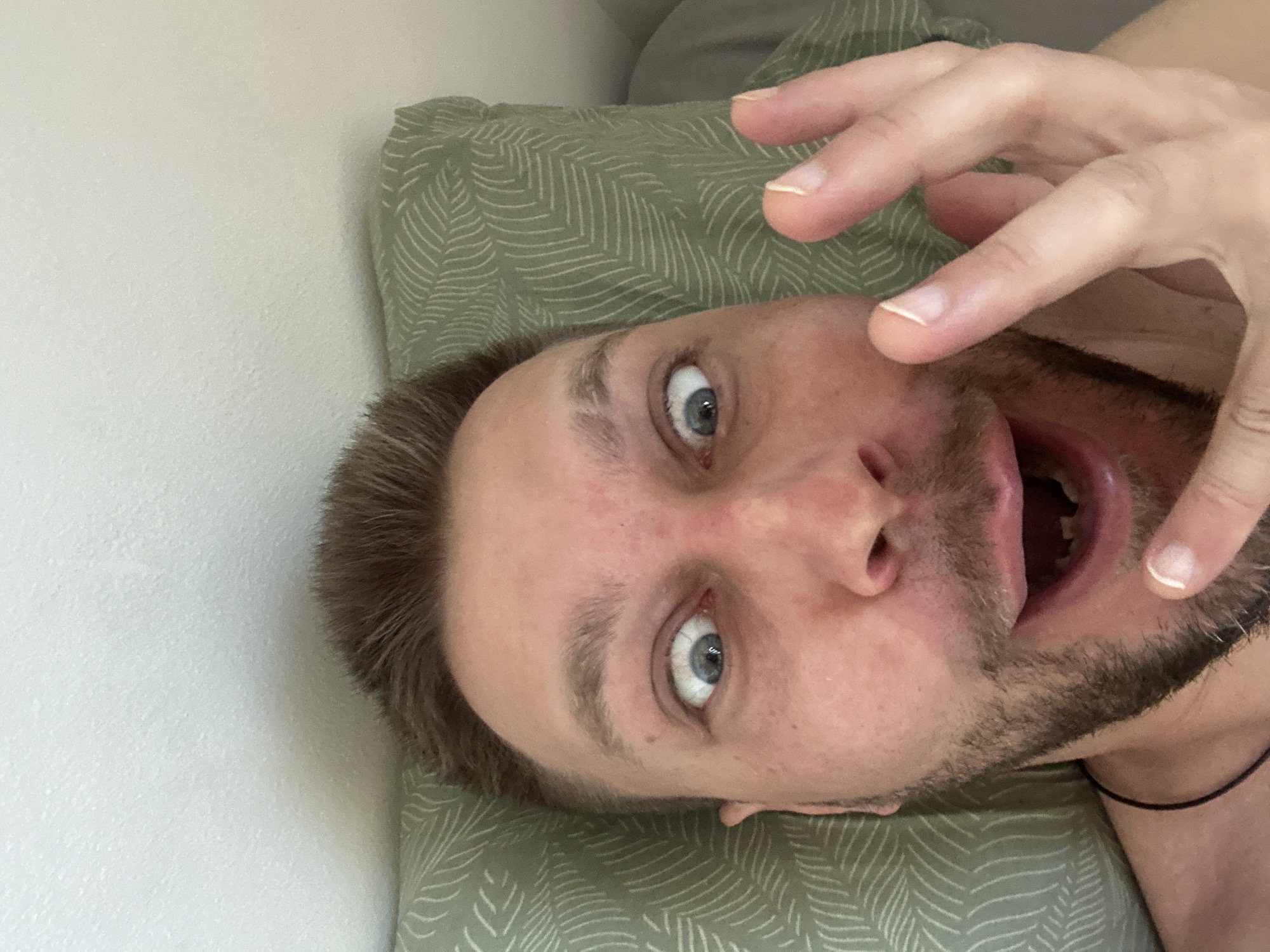 A close-up, first-person perspective selfie shows a man with light brown hair and a beard lying down. His eyes are wide open, and his mouth is open, revealing missing teeth. His left hand is raised, with fingers extended. The man is lying on a green pillow with a leaf pattern and a white wall is visible in the background. The image was taken in Zvezdara Urban Municipality, Serbia.