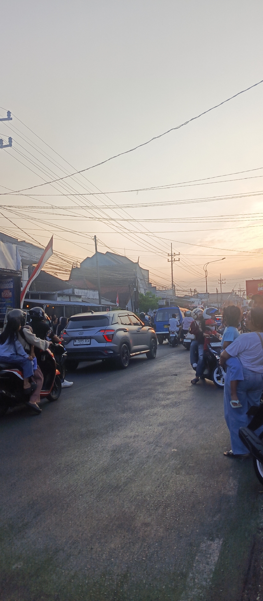 This image depicts a busy street scene in Malang, Indonesia, during what appears to be either dawn or dusk, with the sky exhibiting a warm, hazy glow. The foreground is dominated by a paved road with visible tire tracks and some scattered debris.
Several vehicles are present, including a modern grey SUV with license plate "N 1095 ACH" in the center of the frame. To its left, two individuals are riding on a dark-colored motorcycle, the rider wearing a helmet and the passenger behind them also appearing to be wearing headwear. Further to the left, another dark-colored motorcycle is parked, with a person sitting on it, their face obscured by a helmet.
In the background, multiple other motorcycles and a blue van are visible, suggesting traffic congestion. A crowd of people is gathered on the side of the road, some standing and others on motorcycles. Among the individuals, a woman is carrying a child on her shoulders. A small child, wearing a white shirt, is also seen.
Buildings line the street, with some featuring signage. A large red and white Indonesian flag is prominently displayed, attached to a pole extending from one of the buildings. Another smaller Indonesian flag is visible further down the street. Power lines crisscross the sky, indicating an urban environment. The overall impression is one of a lively, everyday street in a populated area of Malang.