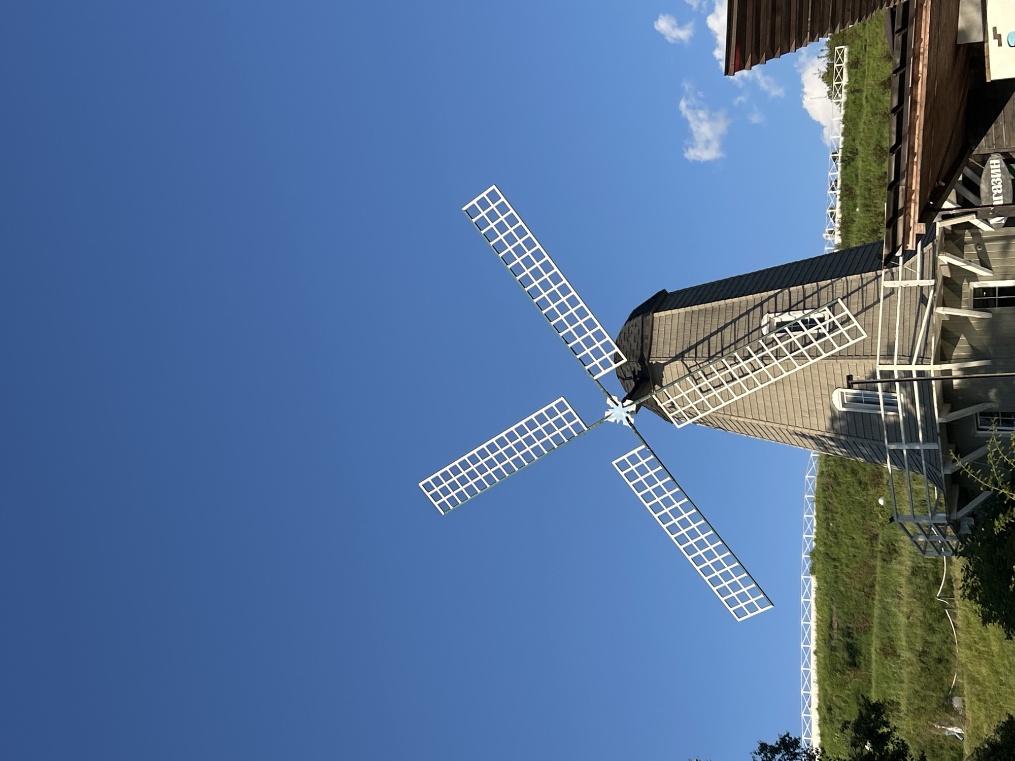 A Dutch-style windmill with white sails stands against a clear blue sky. The windmill is gray with a dark roof. The sails are extended, and each sail is composed of a white grid. Below the windmill, a green grassy hill slopes upwards, with a white fence visible along its crest. In the foreground, there are parts of wooden structures, one with a dark roof and exposed wooden beams, and another section of the windmill with a white railing and windows. The image appears to be taken during the daytime, with ample sunlight casting shadows. The context of the city of Romashkovo, Russia is provided, suggesting this is a specific landmark or attraction within that location.