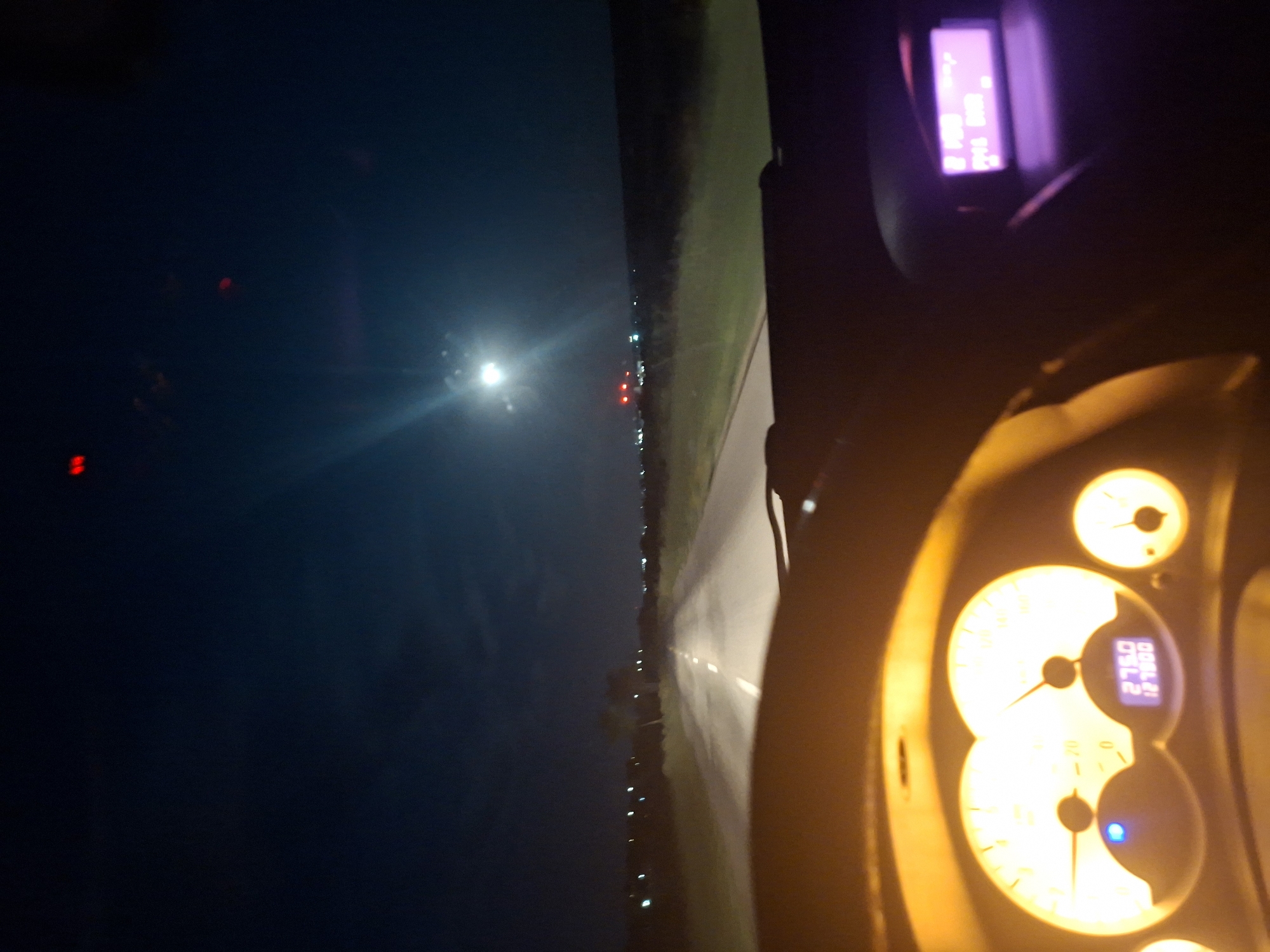 A photograph taken from the driver's seat of a vehicle moving along a road at night. The dashboard gauges are illuminated in orange light and display numbers and indicators. The road stretches ahead, illuminated by the vehicle's headlights, and the sky is dark with some scattered lights visible in the distance. The photograph was taken in Opanets, Bulgaria.