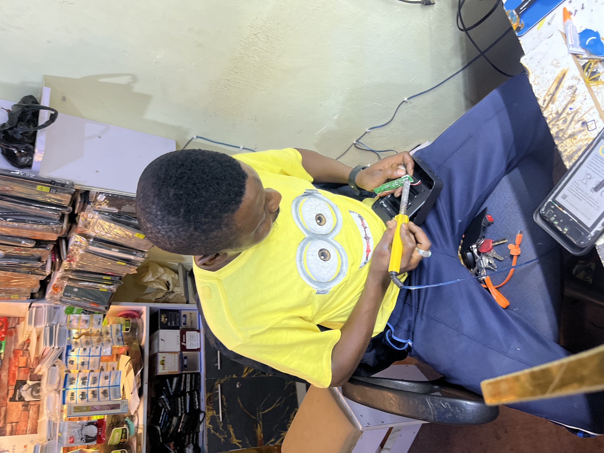 In a cluttered workshop in Bukuru, Nigeria, a man wearing a yellow t-shirt with a Minion graphic and dark blue pants is seated in a black chair. He is intently focused on repairing a circuit board, holding a soldering iron in his right hand and manipulating the board with his left. Tools, including a pair of orange pliers, and a set of keys are scattered on his lap. The background reveals shelves filled with various electronic accessories, phone cases, and other merchandise, suggesting a retail and repair environment. Wires are visible along the wall, and a light-colored wall completes the backdrop.