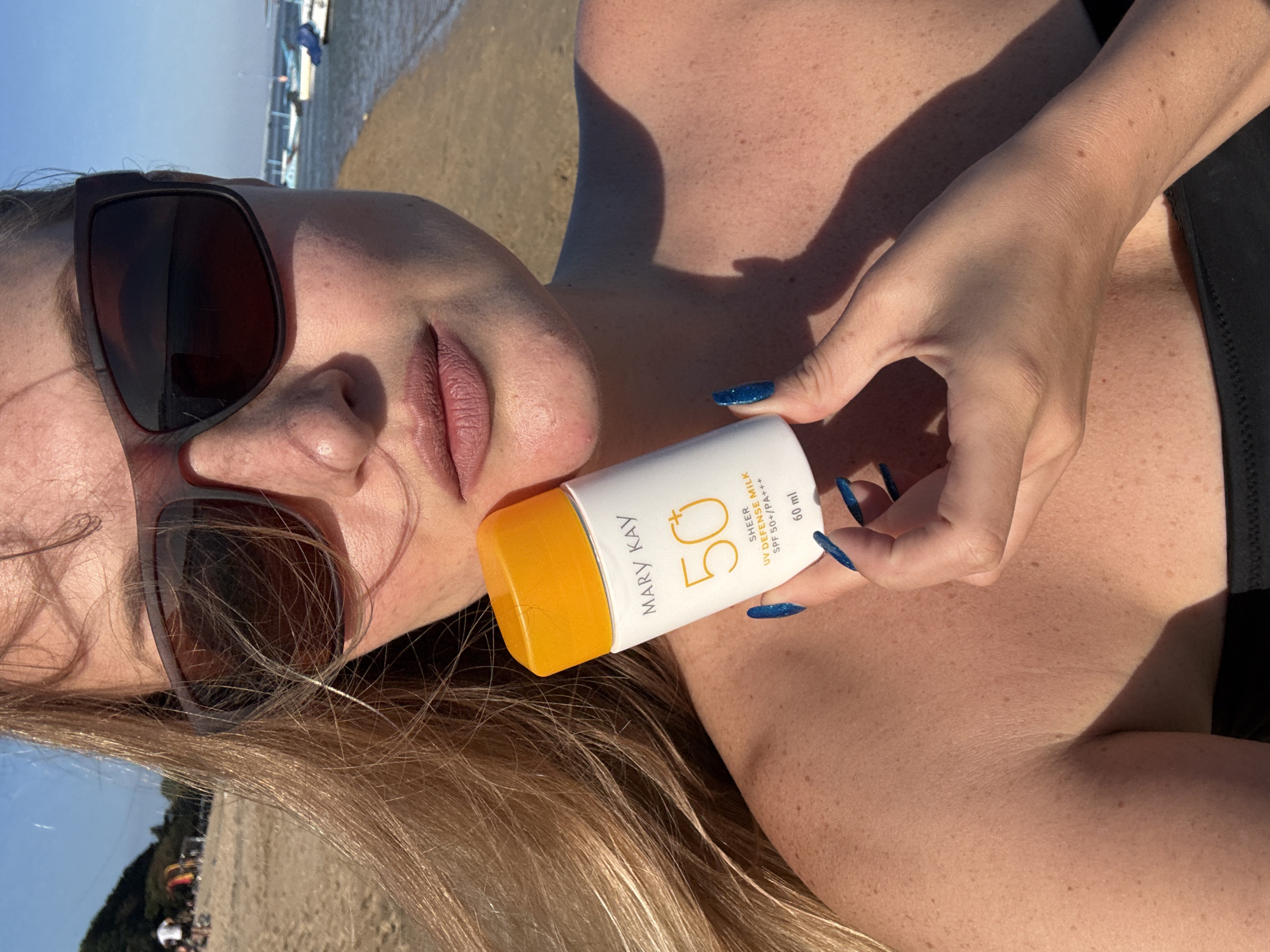 A close-up, head-and-shoulders shot features a fair-skinned woman with blonde hair, wearing large, dark-rimmed sunglasses and a black bikini top. She is holding a white bottle with an orange cap, labeled "MARY KAY 50 SHEER UV DEFENSE MILK SPF 50+/PA+++ 60 ml," in her right hand. Her fingernails are painted a bright blue. The background suggests a beach environment with sand, a body of water, and boats, under a clear blue sky. There are distant figures on the beach and some vegetation on a hill to the left. The image appears to have been taken during daylight hours. The location is the city of Denpasar, Indonesia.