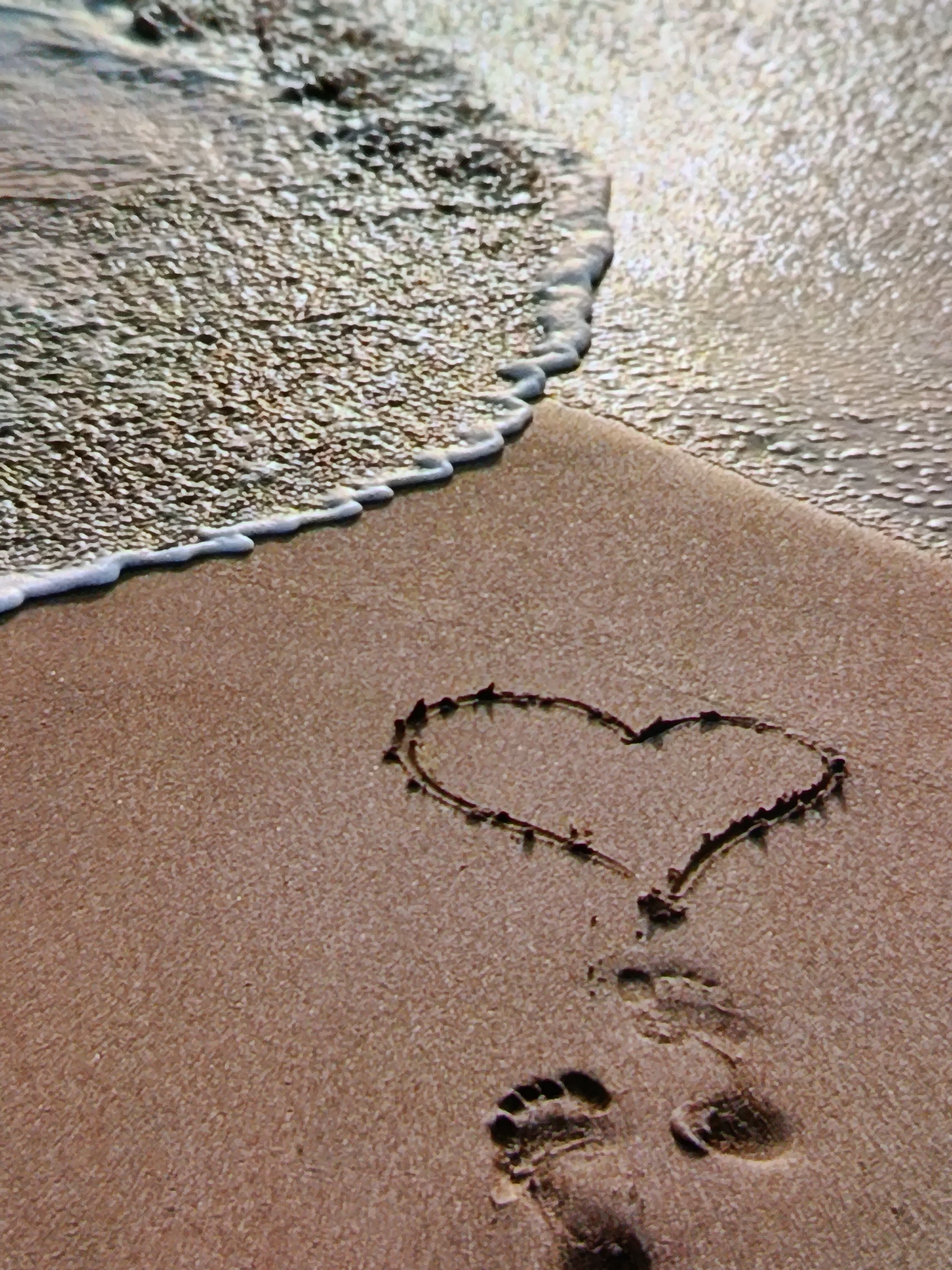 The image depicts a sandy beach with gentle waves lapping at the shore. A heart shape is drawn in the wet sand, accompanied by footprints. The sun glints off the water, creating a shimmering effect. The scene is peaceful and serene. There is no discernible location in Odesa, Ukraine, or any specific details about people, actions, or interactions that would suggest a narrative beyond the visual elements.