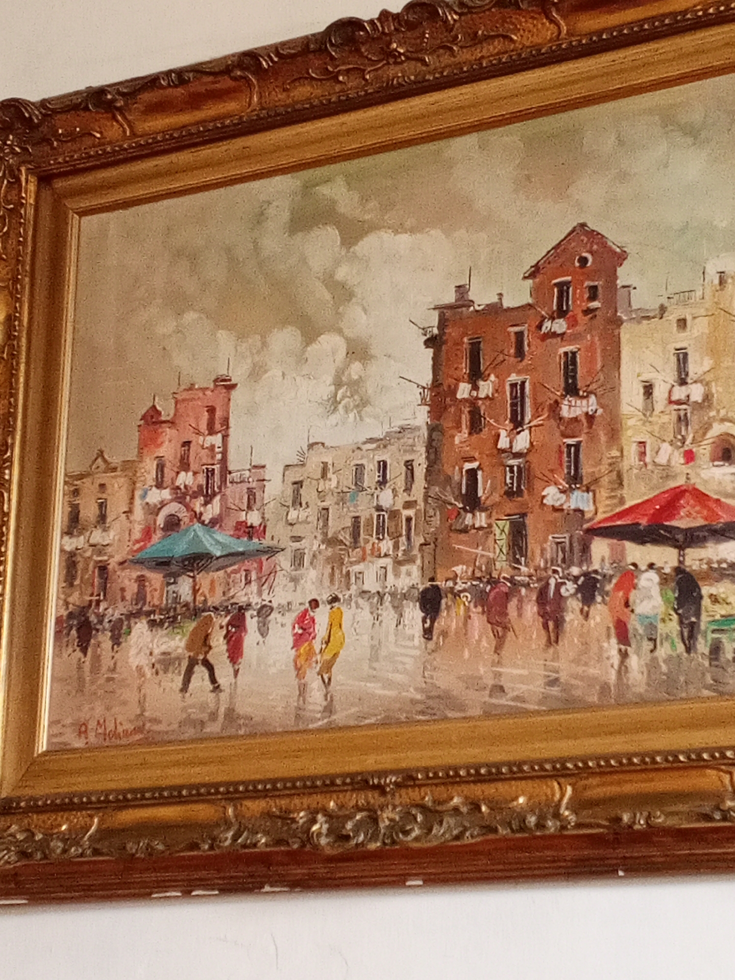A framed painting depicts a bustling city scene. Several multi-story buildings with visible windows and some laundry hanging from them line a square. People of various sizes and colors are walking in the square, with some figures appearing elongated and blurry, perhaps suggesting movement. Two market stalls are visible, one with a red umbrella and the other with a blue umbrella. The sky is overcast with grey and white clouds. The frame is ornate and gold in color. A signature, partially legible as "A. Molina," is located in the lower left corner of the painting. The painting style is impressionistic, with loose brushstrokes and blurred details. The scene does not resemble the city of Tinaquillo, Venezuela.