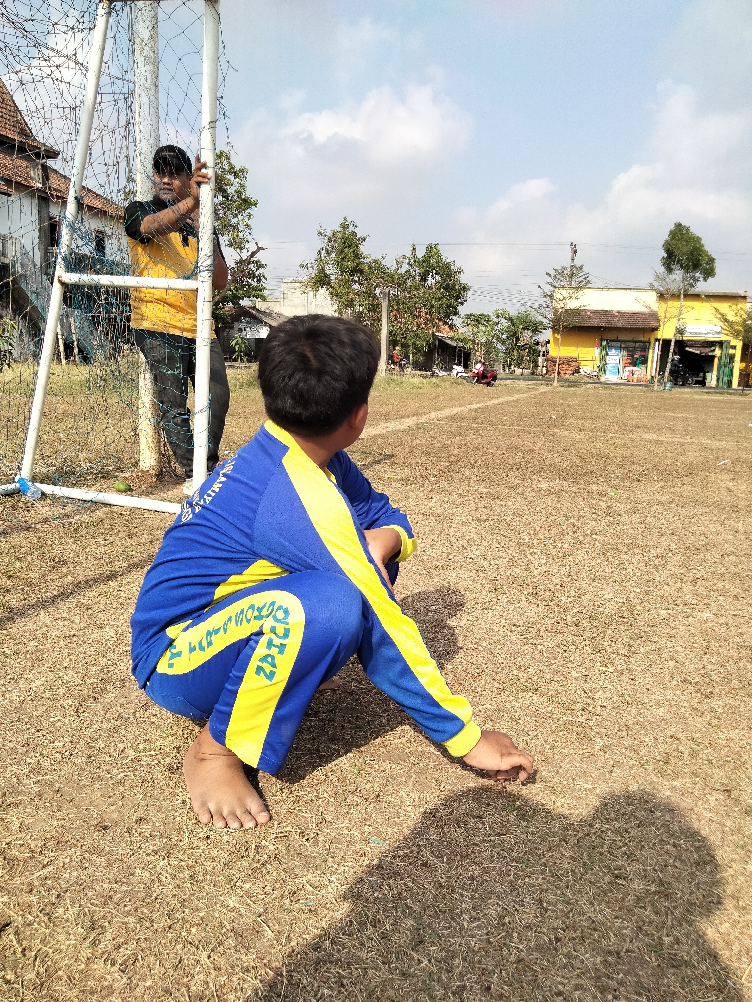 In Pucakwangi, Indonesia, a young boy wearing a blue and yellow tracksuit is crouching on a dry, grassy field. His back is to the camera, and he is looking towards a goalpost. A man in a yellow t-shirt and black pants is standing behind the goalpost, holding onto it. The field appears to be part of a schoolyard or sports complex, with a building and trees visible in the background. The sky is clear and blue with a few scattered clouds. The boy's left foot is bare.