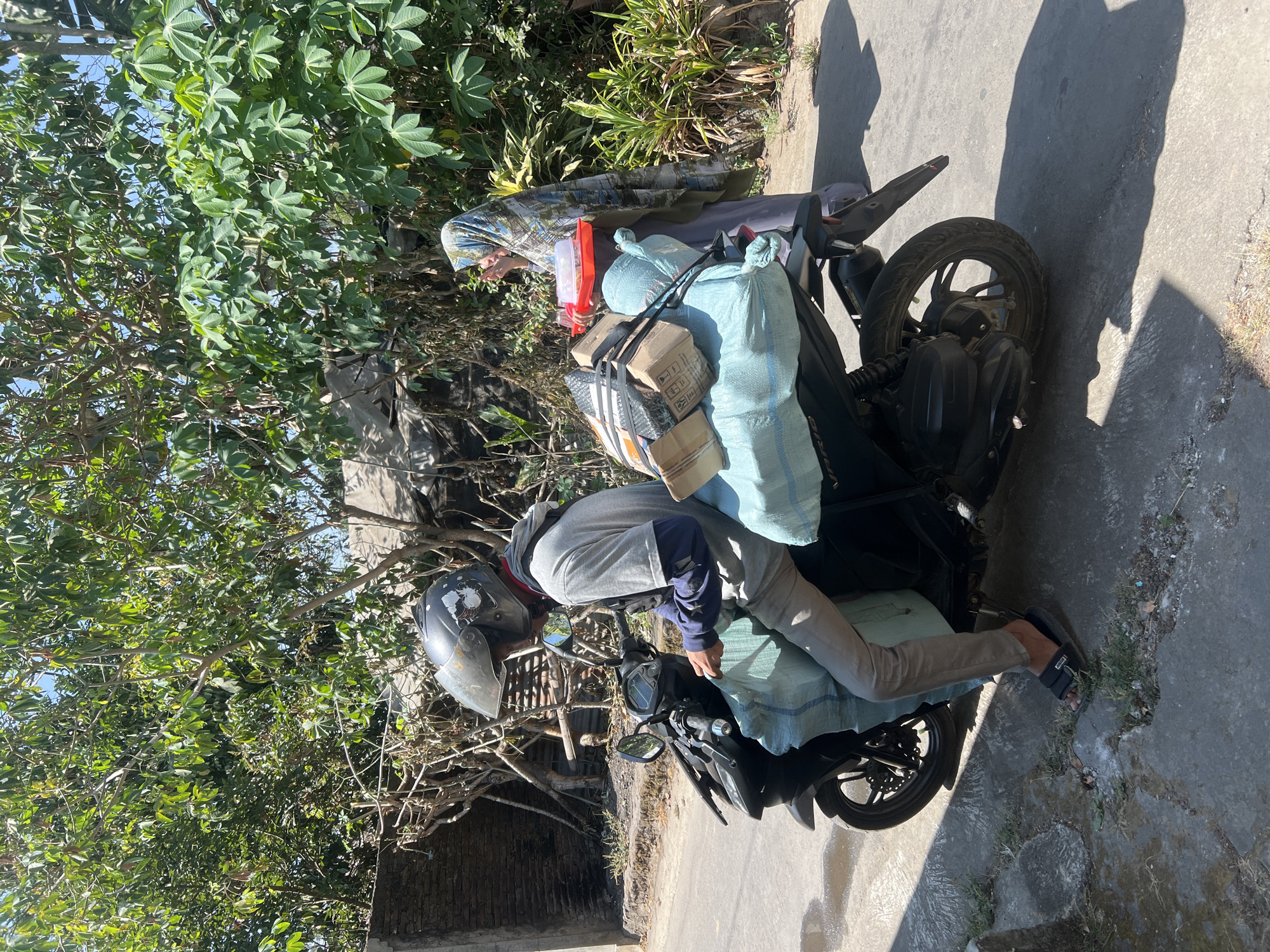 A man wearing a helmet, a gray t-shirt, and khaki pants is sitting on a motorcycle loaded with packages. The motorcycle is parked on the side of a road, which appears to be in a rural area with trees and vegetation. A woman wearing a hijab is standing behind the motorcycle, holding a red tray with items on it. The scene is illuminated by sunlight, casting shadows on the ground. The location is identified as Payaman, Indonesia.