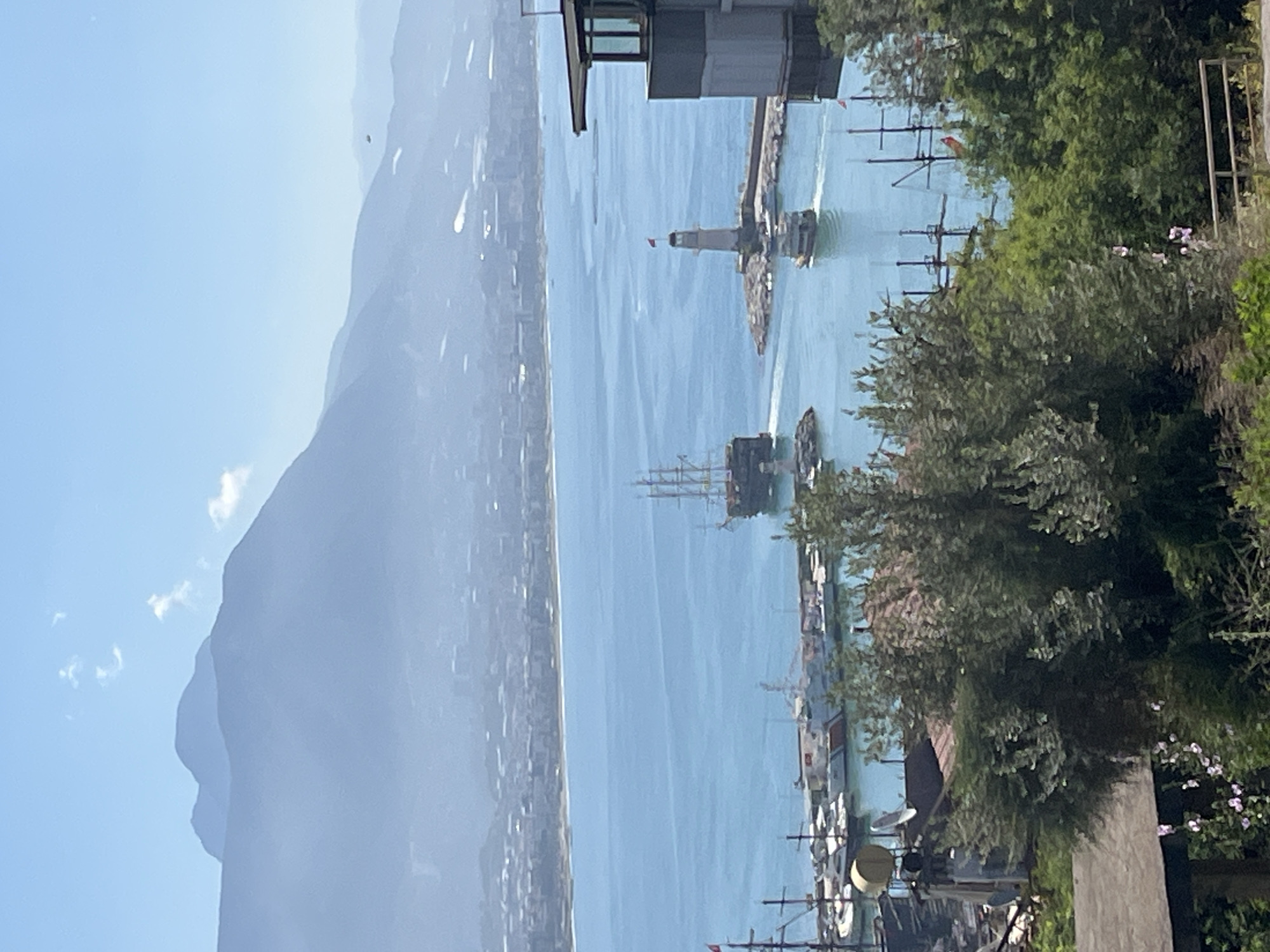 The image shows a cityscape of Alanya, Turkey, with a view of the Mediterranean Sea. In the foreground, there are trees and some buildings. In the mid-ground, there's a lighthouse on a breakwater, several ships in the water, including at least one ship with masts that evoke a historic sailing vessel. There are also several small boats moored along the coast. The background features a hazy coastline with mountains rising in the distance. The sky is clear and light blue. The scene is captured from a high vantage point, possibly from a building or elevated area.