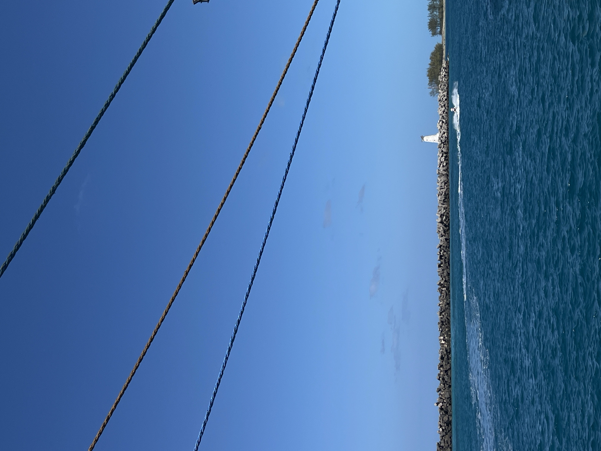 The image shows a body of water, possibly an ocean or large lake, with a rocky shoreline and a small island or peninsula in the distance. A white lighthouse is visible on the island. A personal watercraft is traveling on the water near the shore. In the foreground, there are three ropes or lines hanging down, possibly from a boat or overhead structure. The sky is clear and blue. The location appears to be a coastal area, city unknown.