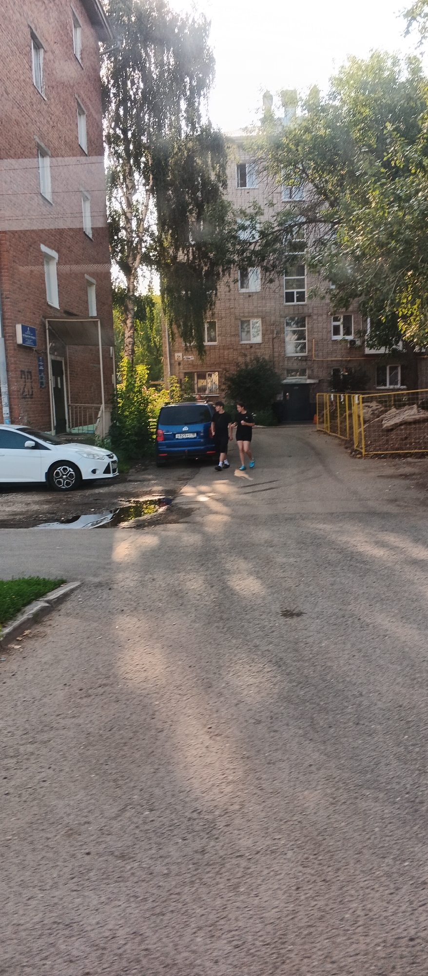 The image depicts a street scene in front of apartment buildings, likely in Izhevsk, Russia. On the left, a white car is parked. To the right of the car, a blue minivan is parked, and two young individuals, one appearing to be wearing dark clothing and the other a dark t-shirt and shorts, are walking away from the minivan. Behind the minivan, a yellow metal fence encloses an area with debris. Tall trees with green foliage are visible, casting shadows on the paved ground. The buildings are multi-story brick structures with visible windows and balconies. The scene appears to be captured during daylight, possibly in the late afternoon, given the sunlight and shadows. A street sign with Cyrillic lettering is affixed to one of the buildings. A puddle of water is present on the asphalt near the white car.