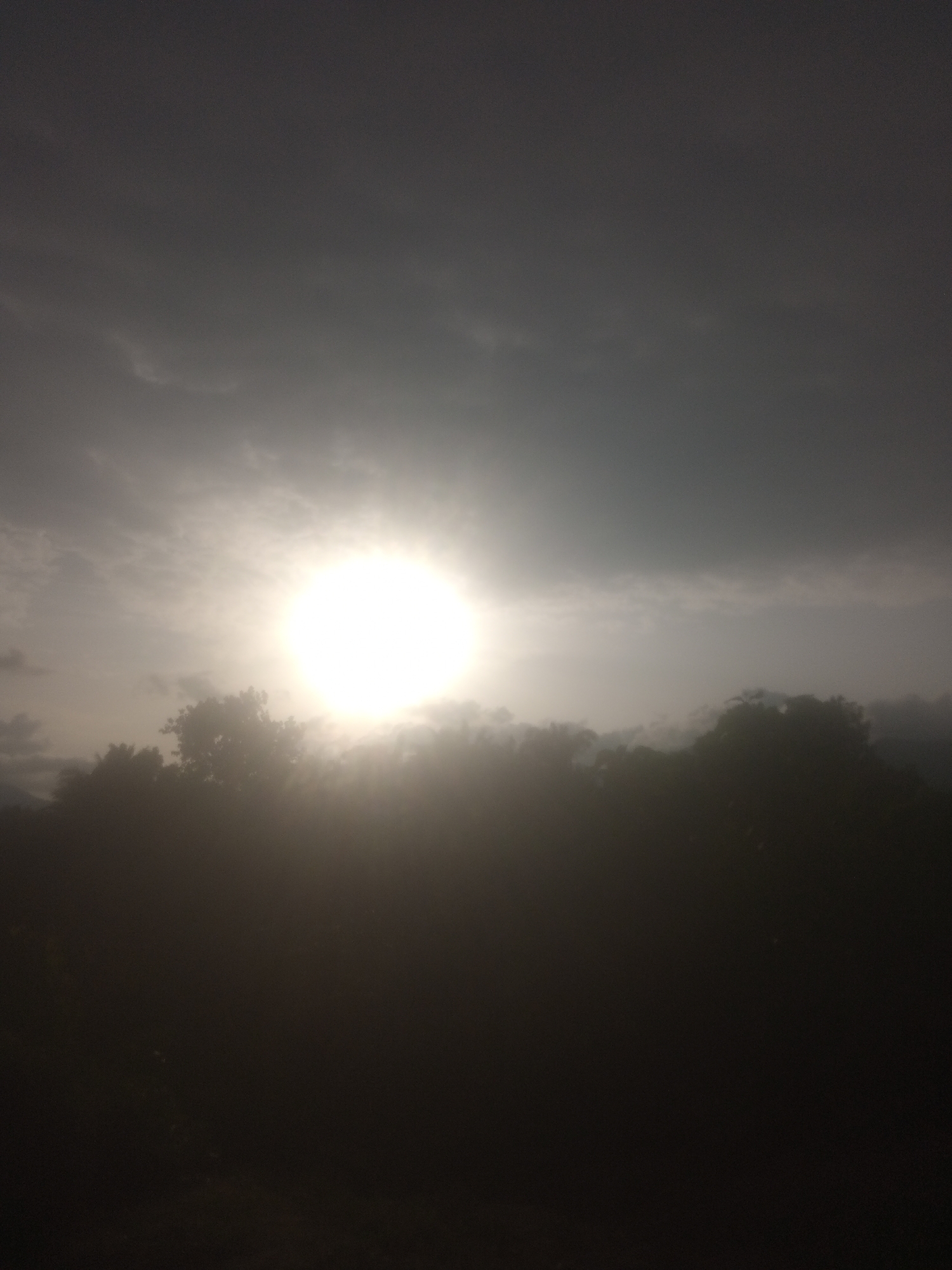 This media file captures a hazy view of the sun shining brightly through clouds, casting a glow over a silhouetted line of trees. The sun is the central subject, appearing as a brilliant white orb with rays of light emanating outwards, suggesting it is either rising or setting. The sky is filled with varying shades of gray and white clouds, with some darker areas indicating denser cloud cover.
The lower portion of the image is dominated by the dark silhouettes of trees, creating a natural horizon. The overall scene is somewhat obscured by atmospheric haze or mist, which softens the details of the foliage and creates a dreamy, diffused effect. The light intensity and the presence of clouds suggest a time of day when the sun is low in the sky, likely dawn or dusk. The location is Serti, Nigeria, and the scene evokes a sense of natural beauty and tranquility. There is no visible text in the image.