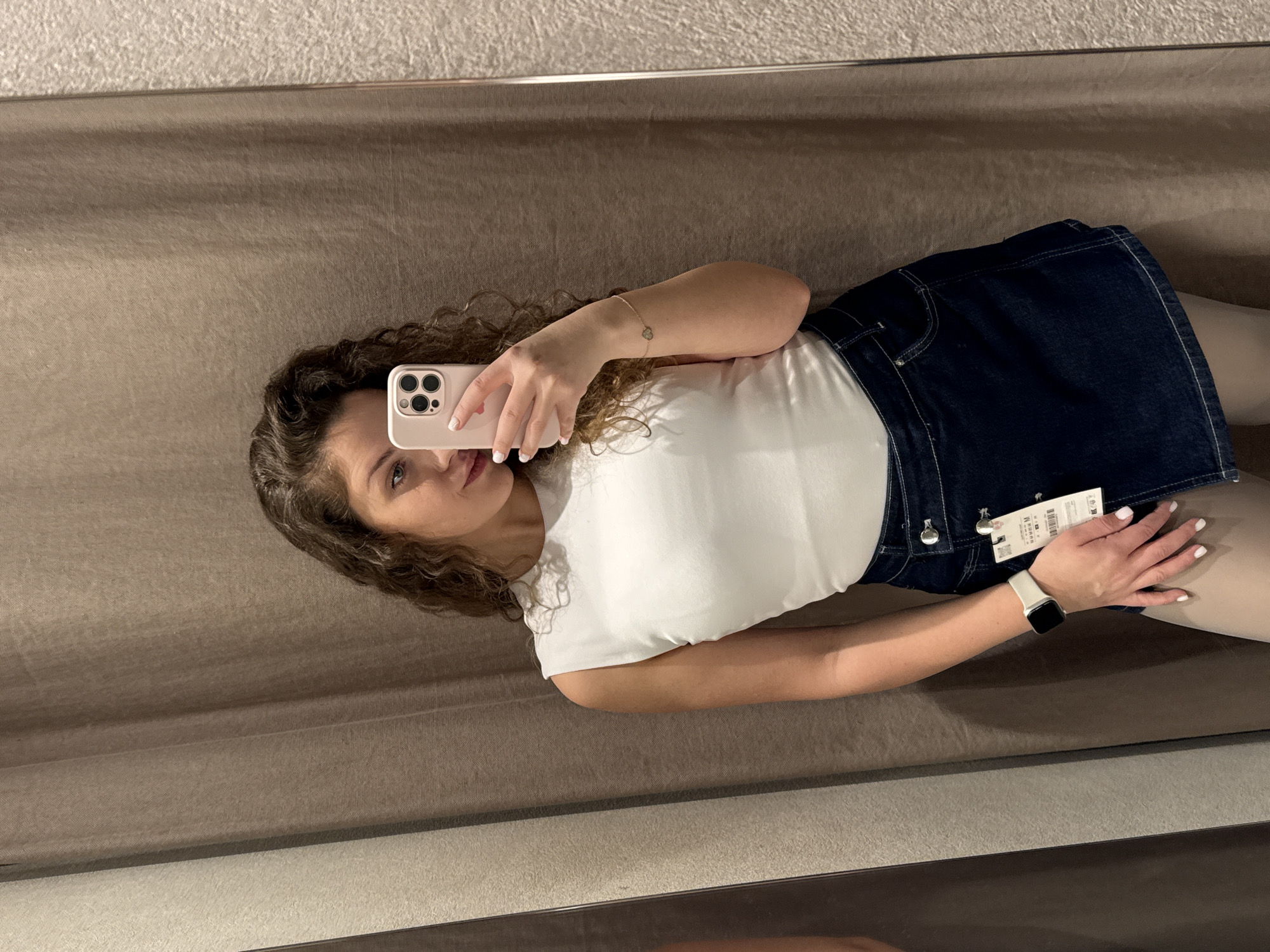 A woman is taking a selfie in what appears to be a dressing room. She has curly brown hair and is wearing a white t-shirt and a dark blue denim skirt with a price tag still attached. She's holding a pink smartphone up to take the photo. The background is a plain tan wall, likely the inside of a fitting room. The scene suggests a retail environment, possibly a clothing store. There is no indication of the location being Sofia, Bulgaria or anywhere else.