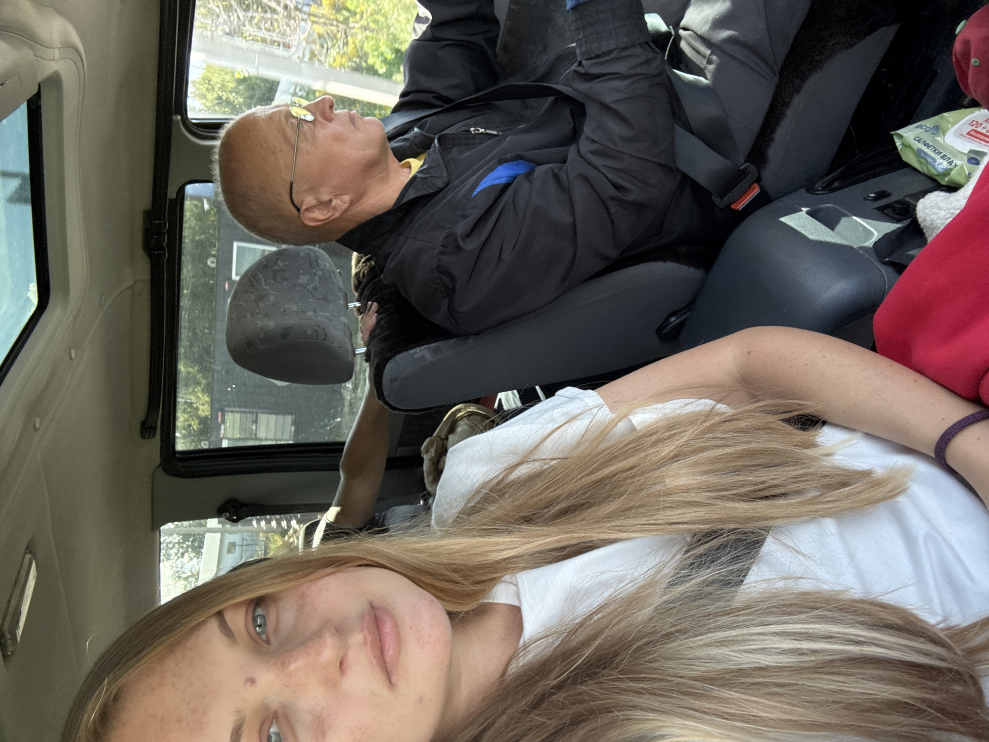 In the city of Razvitie, Russia, a young woman with long blonde hair and freckles is taking a selfie in the passenger seat of a car. She is wearing a white t-shirt. To her right, an older man with glasses is driving. He is wearing a black jacket over a yellow shirt. The car has a sunroof and the interior is visible, along with a glimpse of the outside environment. The scene appears to be during the daytime.