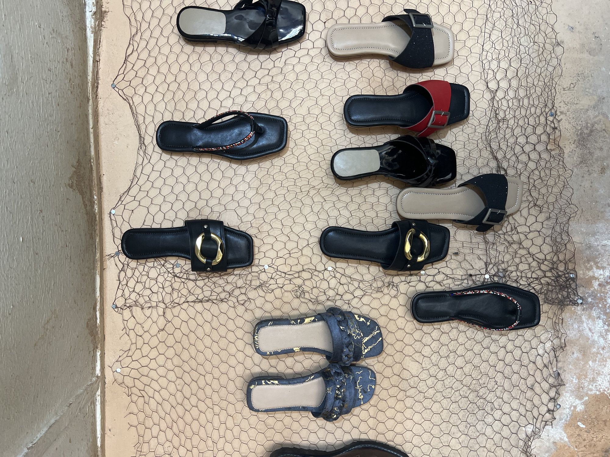 The image depicts a display of sandals hanging on a wall in Jos, Nigeria. The sandals are arranged in rows and columns on a wire mesh attached to a wall. The wall appears to be painted in a light brown color. The sandals vary in style, color, and material. They include black leather sandals, sandals with gold buckles, and sandals with red straps. The wire mesh is secured to the wall with nails. The environment is basic.