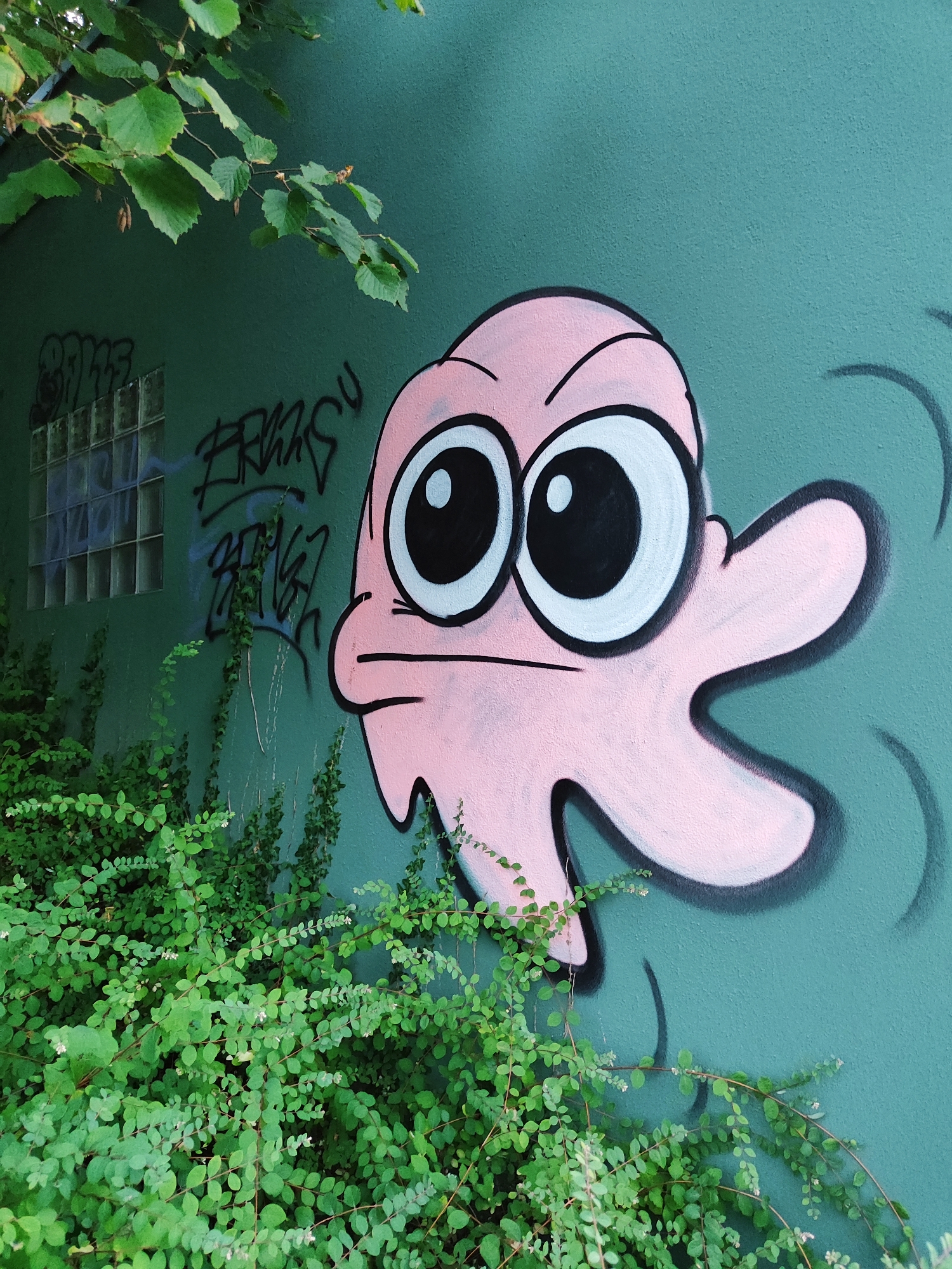 The image displays a graffiti artwork of a pink, cartoonish creature with large, expressive eyes painted on a teal-colored wall. The creature has a round head with a wide mouth and what appear to be three appendages extending from its body. The artwork is located in Offenburg, Germany, near a building with a glass block window. Lush green foliage, likely ivy or a similar climbing plant, covers the lower portion of the wall and extends onto the ground in front of it, partially obscuring the graffiti. There are also some visible tags in black spray paint on the wall above the greenery and to the left of the main artwork. The overall scene suggests an urban outdoor setting with elements of nature intertwined with street art.
