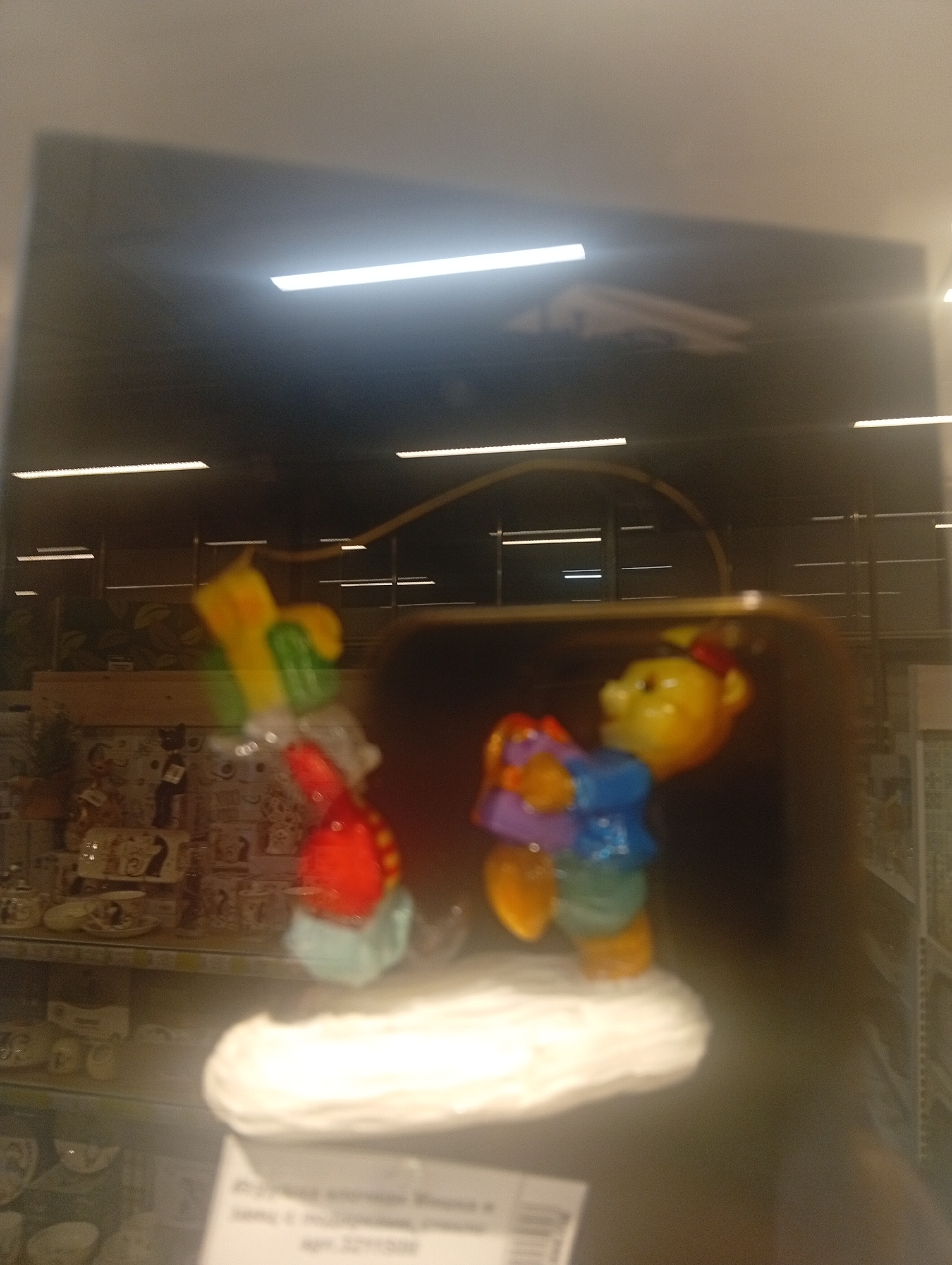 This is a blurry, overexposed, wide-angle photograph taken indoors, likely in a retail environment, possibly a department store or a supermarket. The image is dominated by reflections, making it difficult to discern specific details. In the foreground, there are two blurred figurines, one yellow and red with a green element, and another yellow-faced figurine wearing a blue and purple outfit. These figurines appear to be on display. Behind the figurines, there are shelves stocked with various items, some of which resemble kitchenware or decorative objects, also blurred. The background shows a dark ceiling with bright horizontal fluorescent lights. The overall impression is that of a dimly lit store interior, with the bright lights creating glare and reflections. The text at the bottom of the image appears to be in Russian, but is unreadable due to the blur and reflections. This image is not sufficient to determine the exact location in Pushkino, Russia.