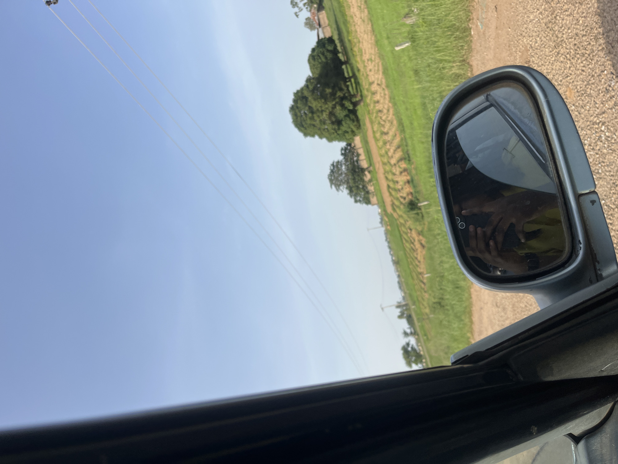 The image depicts a view from inside a vehicle, looking out towards a rural landscape. A side-view mirror is prominently featured, reflecting the image of a person holding a phone, presumably taking the photo, and the interior of the car. Outside the vehicle, a dirt road leads to a field with rows of crops, likely agricultural land. Several trees are scattered across the landscape, and in the distance, utility poles and power lines are visible against a clear blue sky. The setting appears to be a sunny day in a rural area of Ladura Foron, Nigeria.