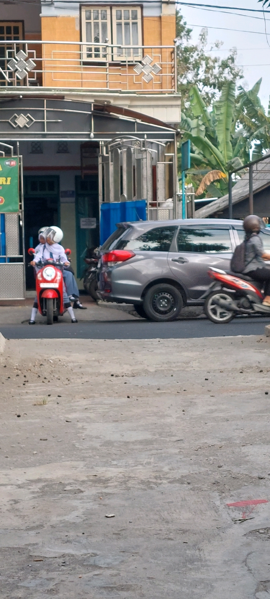 The image shows a street scene in a city. In the foreground, a gray asphalt road with patches of concrete is visible. On the right side of the road, there is a red marking with a stand. In the middle ground, a gray SUV is parked on the road, with its rear end facing the camera. To the left of the SUV, a red scooter is ridden by two individuals wearing white uniforms and helmets. Behind the scooter and SUV, there is a building with an orange facade and a balcony with decorative railings. Adjacent to the building, lush green banana trees are present. A motorcycle ridden by a person with a black backpack is moving to the right, next to the SUV. The environment appears to be daytime, with a cloudy sky.